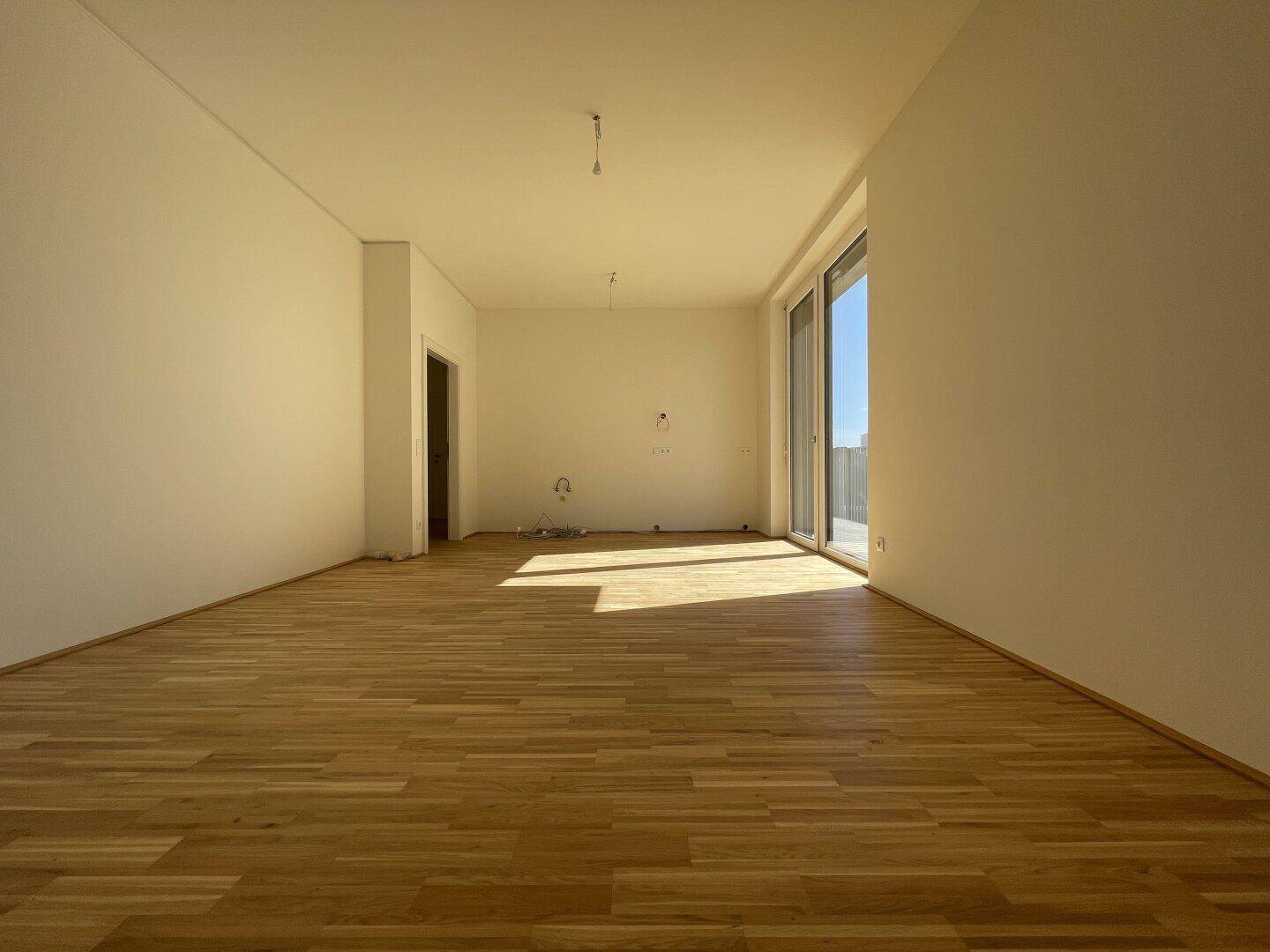 Geräumiges, leeres Wohnzimmer mit hellem Parkettboden und großen Fenstern, die viel Licht hereinlassen.
