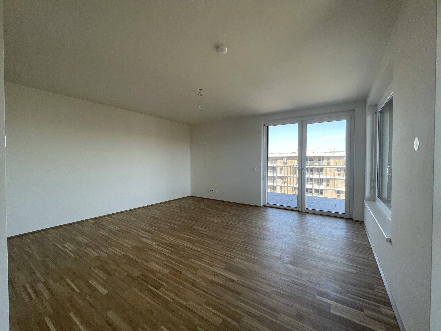 Helles, leeres Wohnzimmer mit Parkettboden und Zugang zu einem Balkon mit Stadtblick.