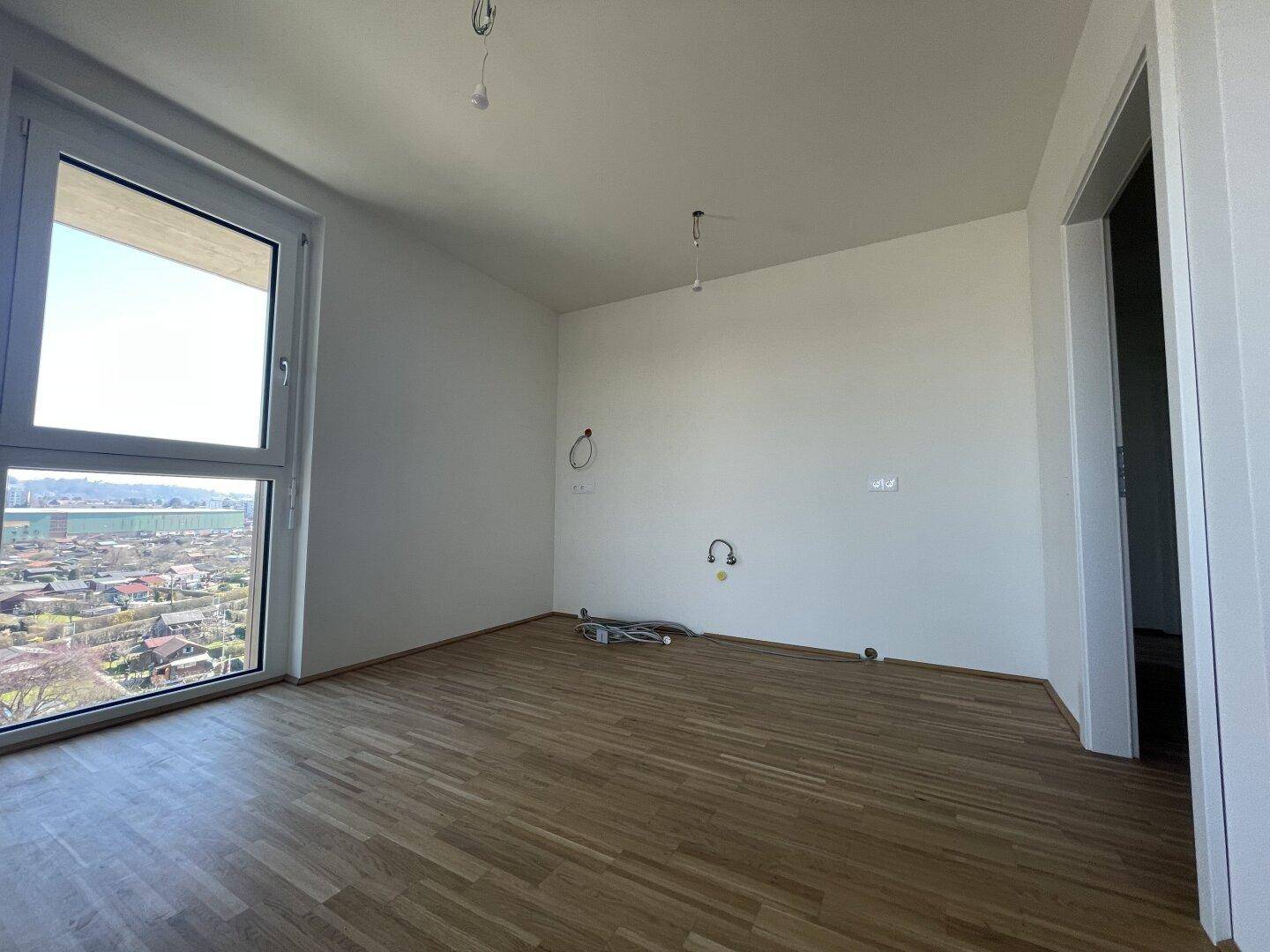 Leerer Küchenraum mit Parkettboden, großem Fenster und vorbereiteten Anschlüssen für die Kücheninstallation.