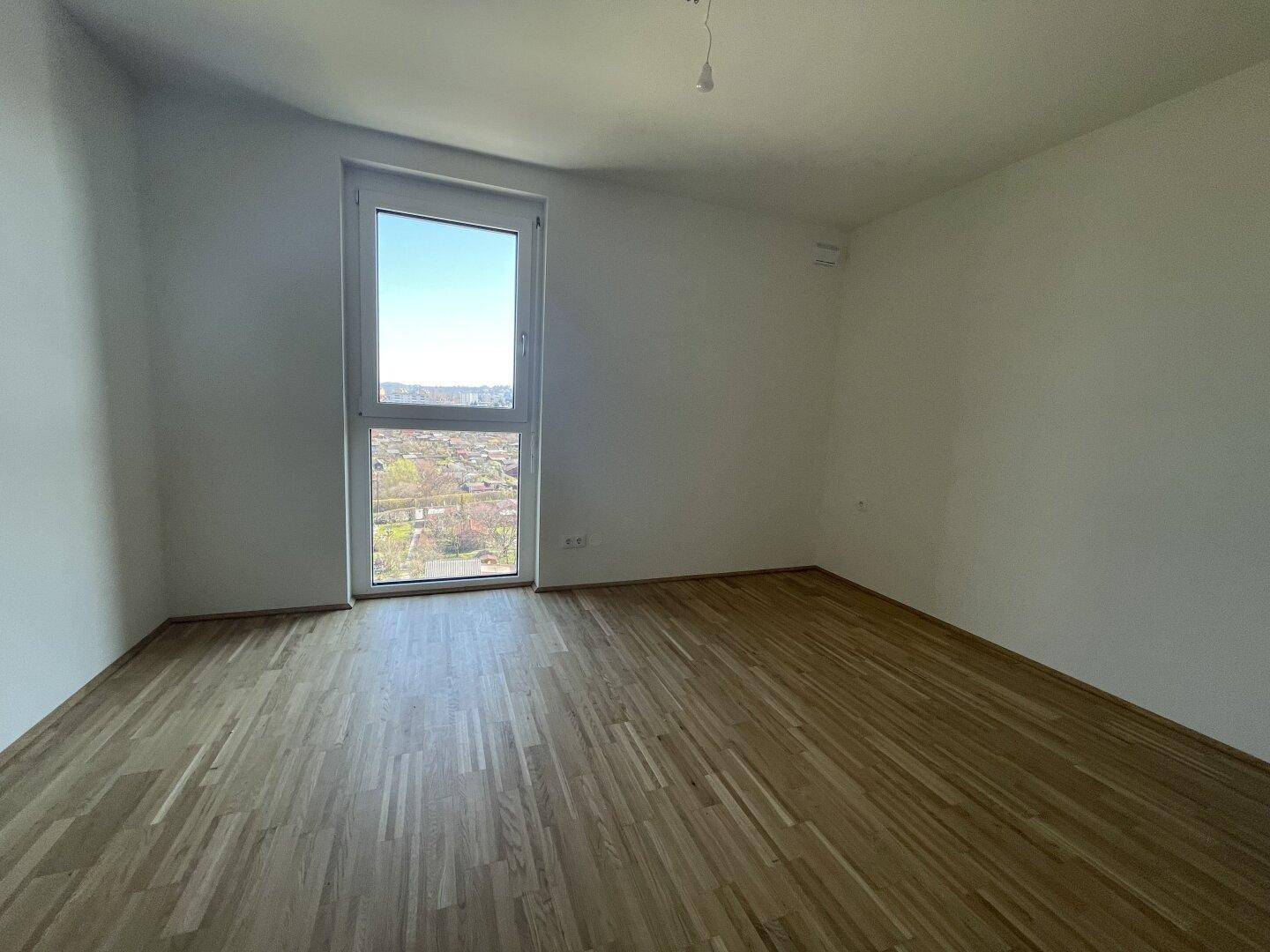 Leeres Schlafzimmer mit Parkettboden und einem großen Fenster, das viel Tageslicht hereinlässt.