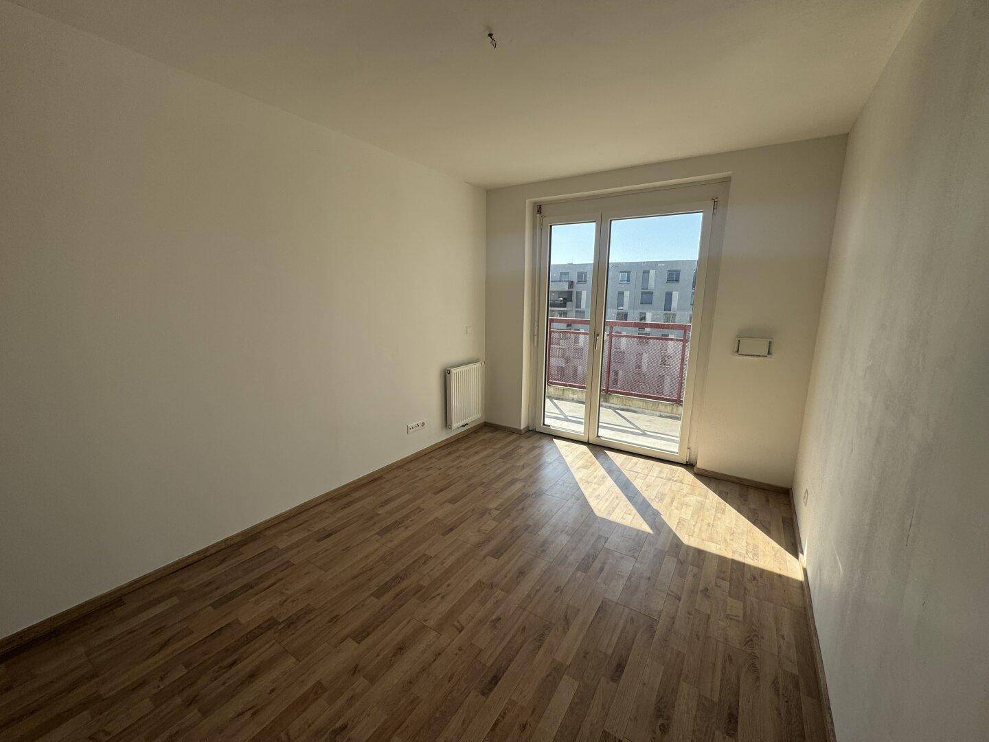 Helles Schlafzimmer mit Laminatboden und großem Fenster, das viel Sonnenlicht hereinlässt.