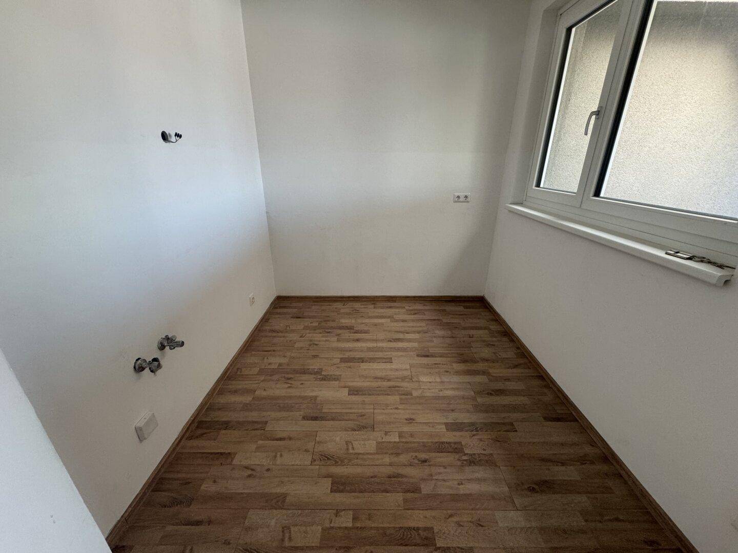 Leerer Raum mit Laminatboden und Fenster, vorbereitet für Installationen mit sichtbaren Anschlüssen.