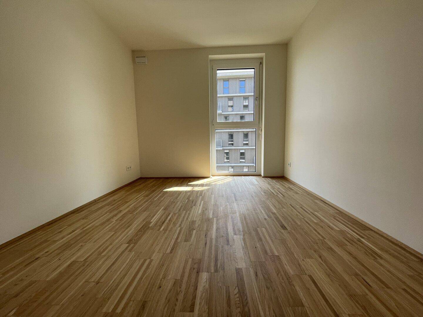 Leeres Schlafzimmer mit hellem Parkettboden und großem Fenster, das viel Tageslicht hereinlässt.