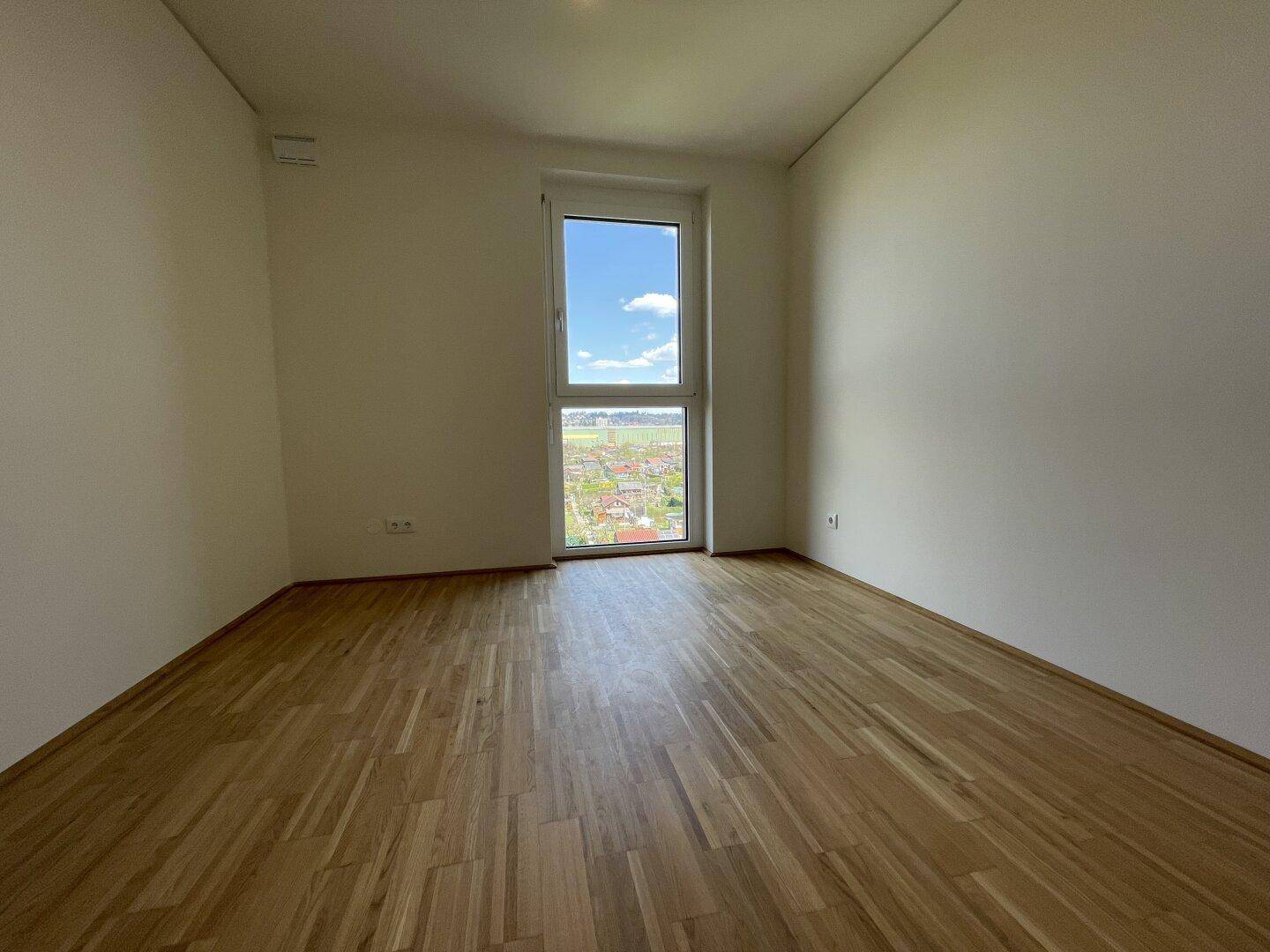 Helles, unmöbliertes Schlafzimmer mit Parkettboden und großem Fenster mit Ausblick.