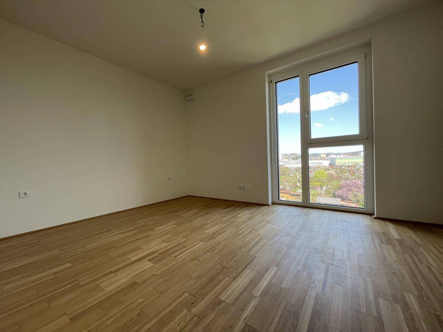 Helles Schlafzimmer mit Parkettboden und großem Fenster, das viel Tageslicht hereinlässt.
