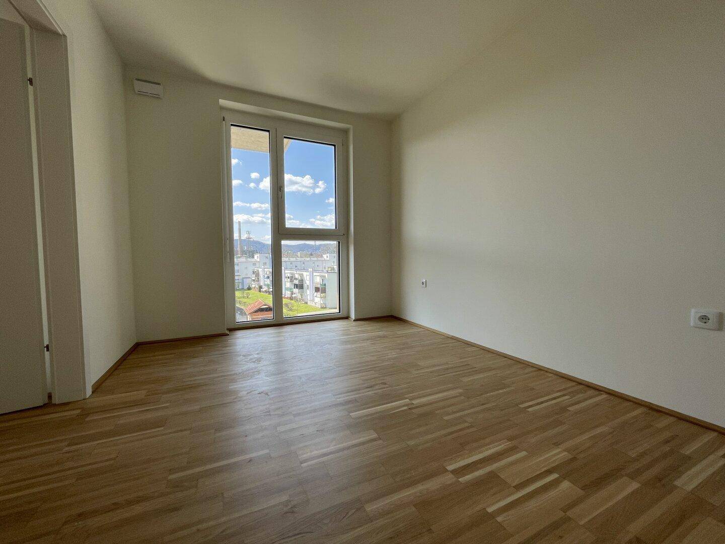Helles Zimmer mit Parkettboden und großem Fenster, bietet viel Tageslicht und einen Ausblick.