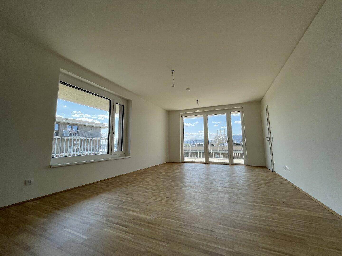 Helles Wohnzimmer mit Parkettboden, weißen Wänden und großen Fenstern mit Balkonzugang.