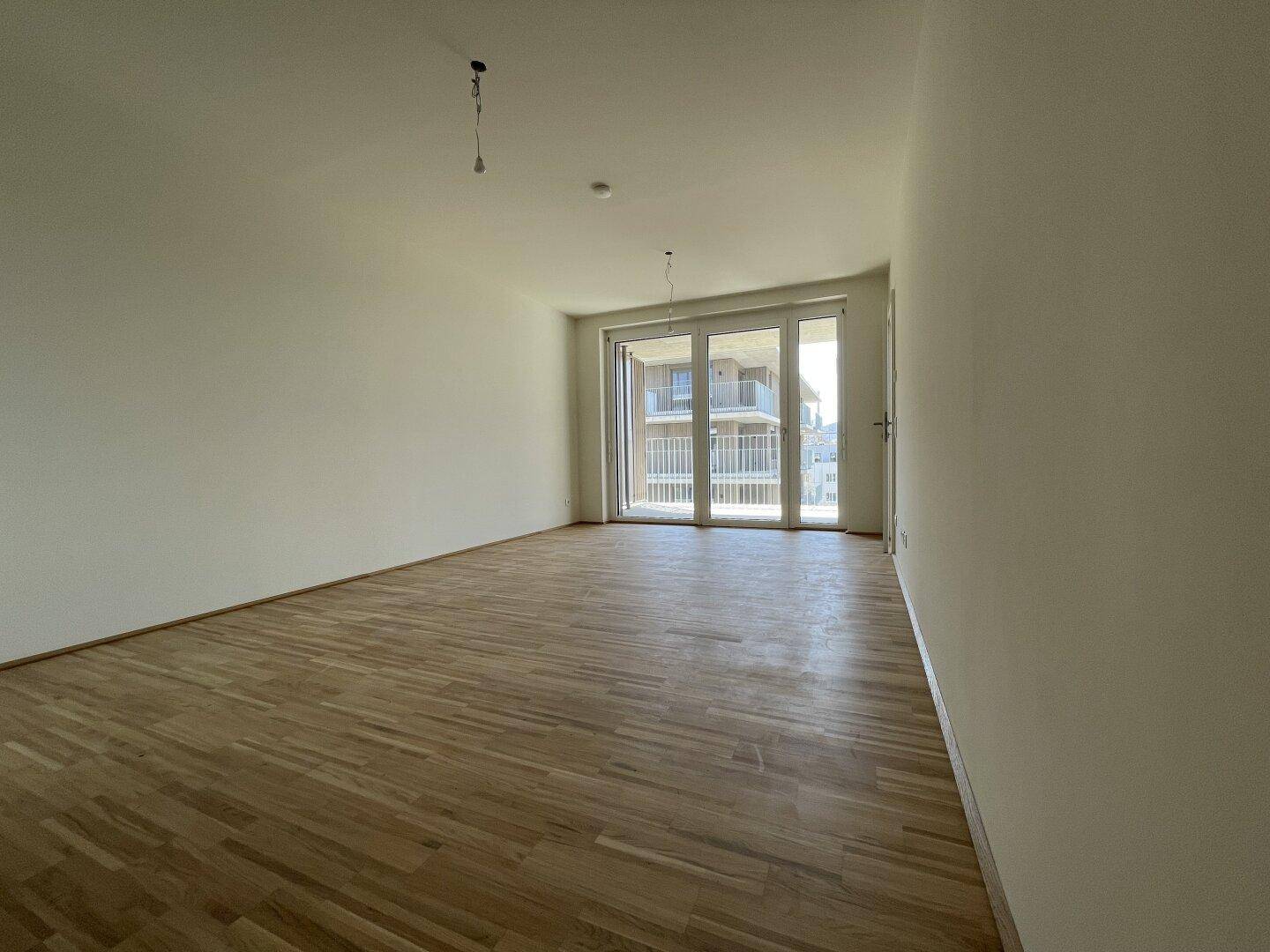 Helles, leeres Wohnzimmer mit Parkettboden und Zugang zu einem Balkon.
