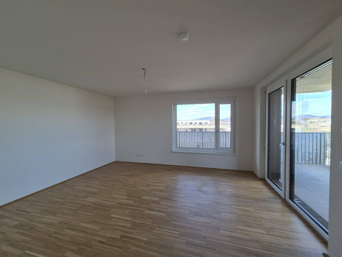 Helles, leeres Wohnzimmer mit Parkettboden und Zugang zu einem großen Balkon.