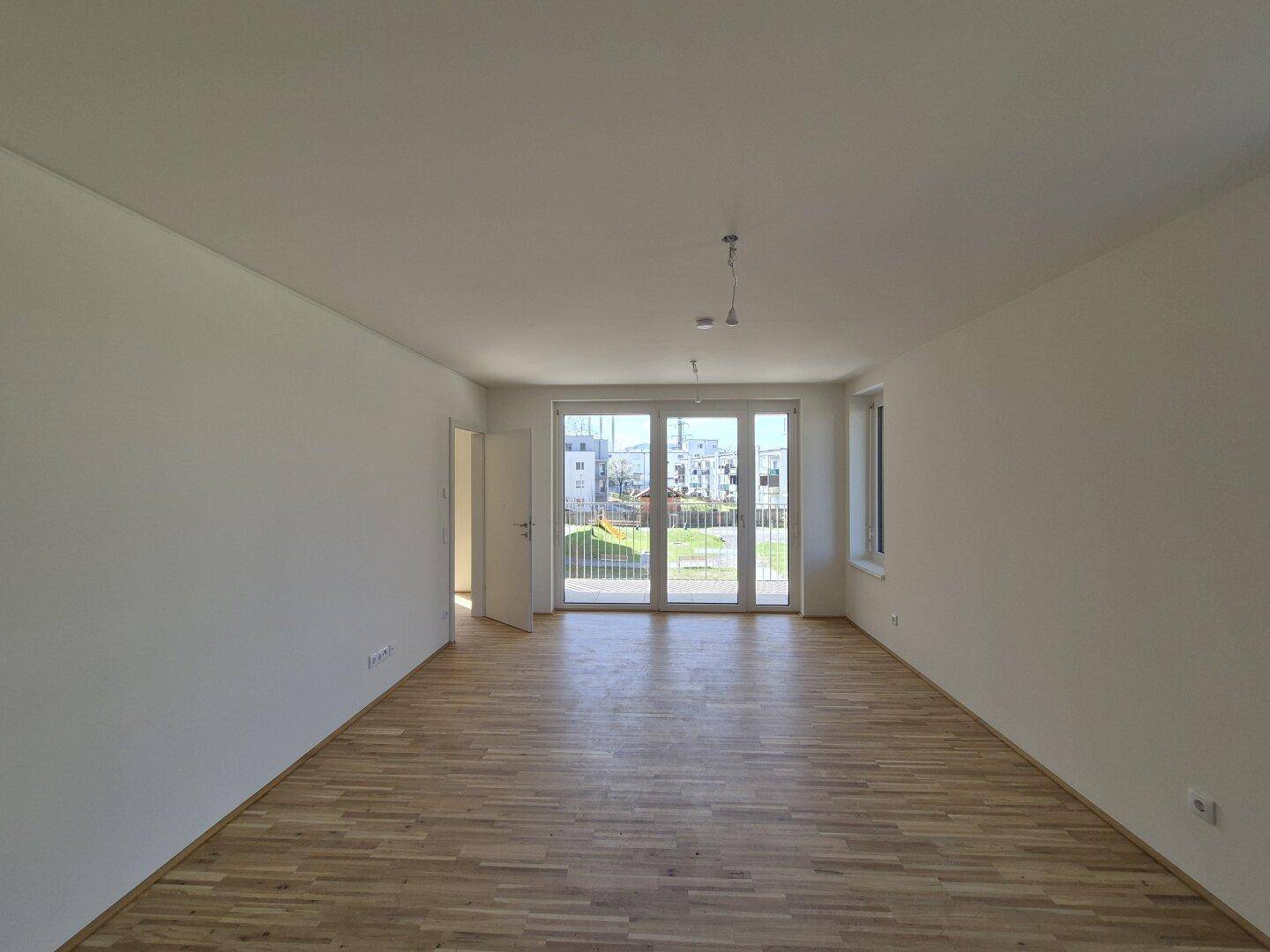 Helles, leeres Wohnzimmer mit Parkettboden und Zugang zu einem Balkon oder Garten.