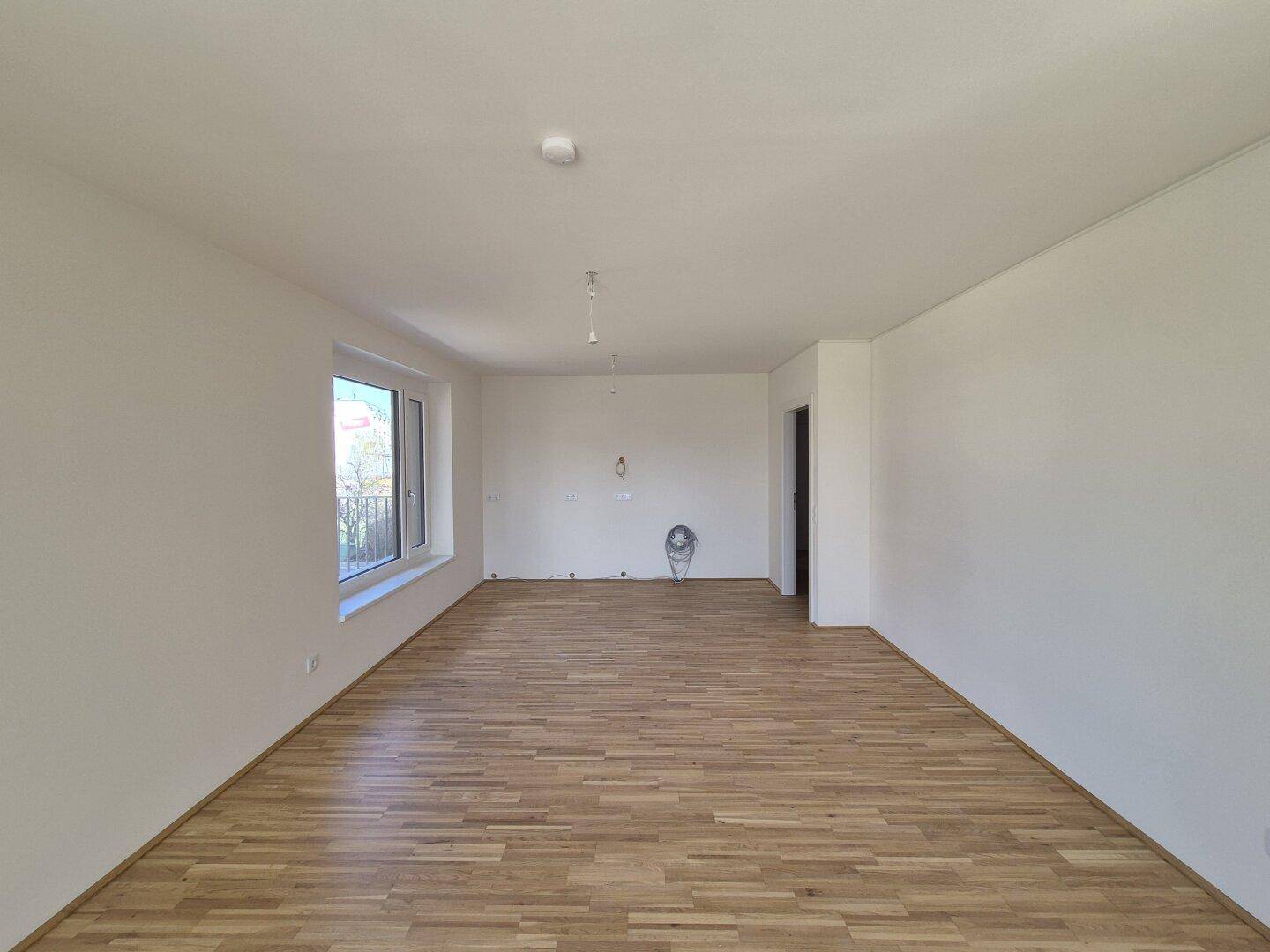 Geräumiges, unmöbliertes Wohnzimmer mit hellem Parkettboden und großen Fenstern.