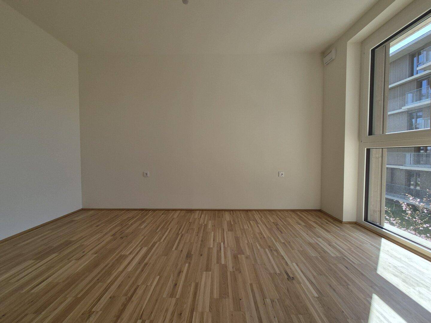Helles Schlafzimmer mit Parkettboden und großem Fenster, das viel Tageslicht hereinlässt.