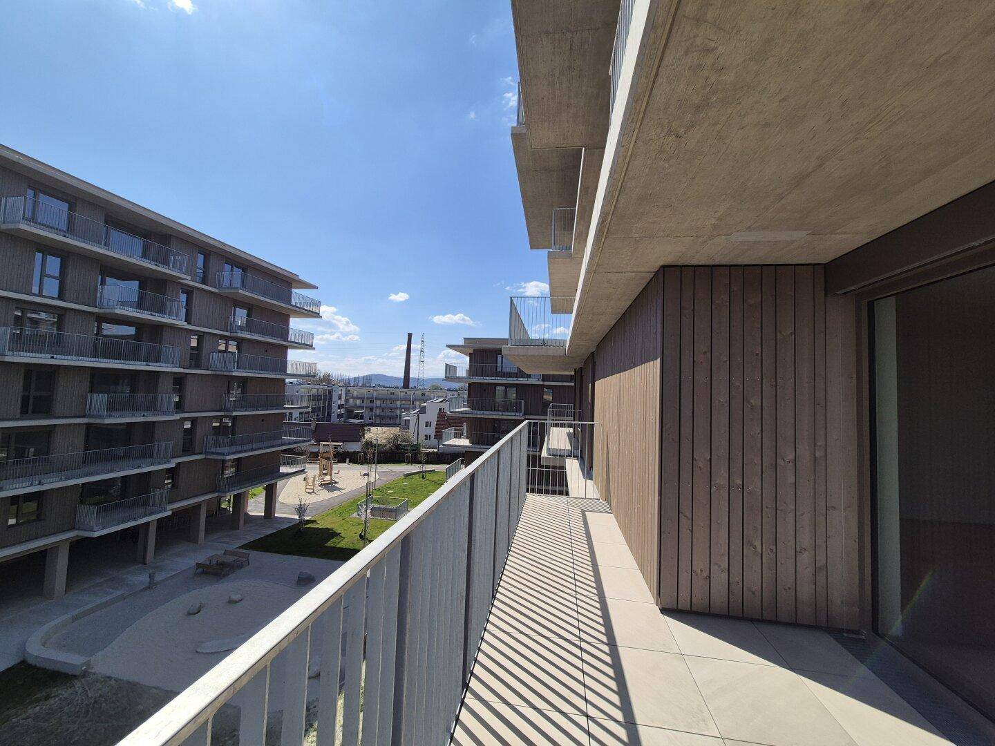 Langer Balkon mit Holzwandverkleidung und Blick auf den Innenhof und weitere Neubauten.