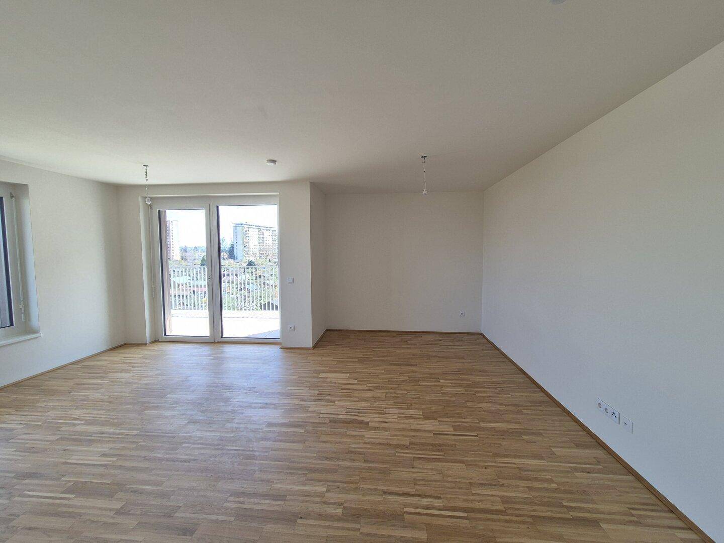 Helles, leeres Wohnzimmer mit Parkettboden und Zugang zu einem Balkon mit Stadtblick.