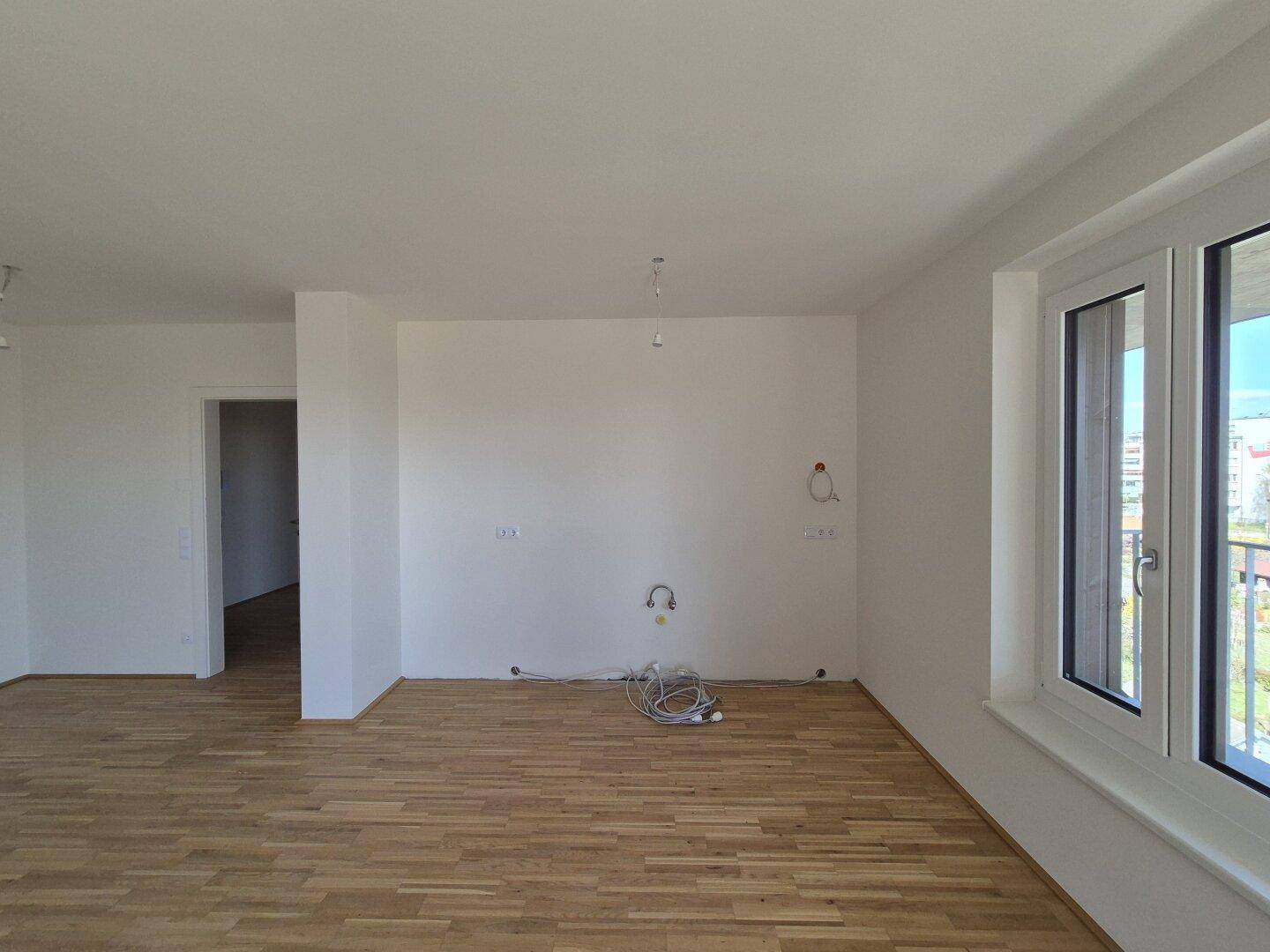 Heller Raum mit Parkettboden, vorbereitet für eine Küche, mit Anschlussmöglichkeiten und Fenster.