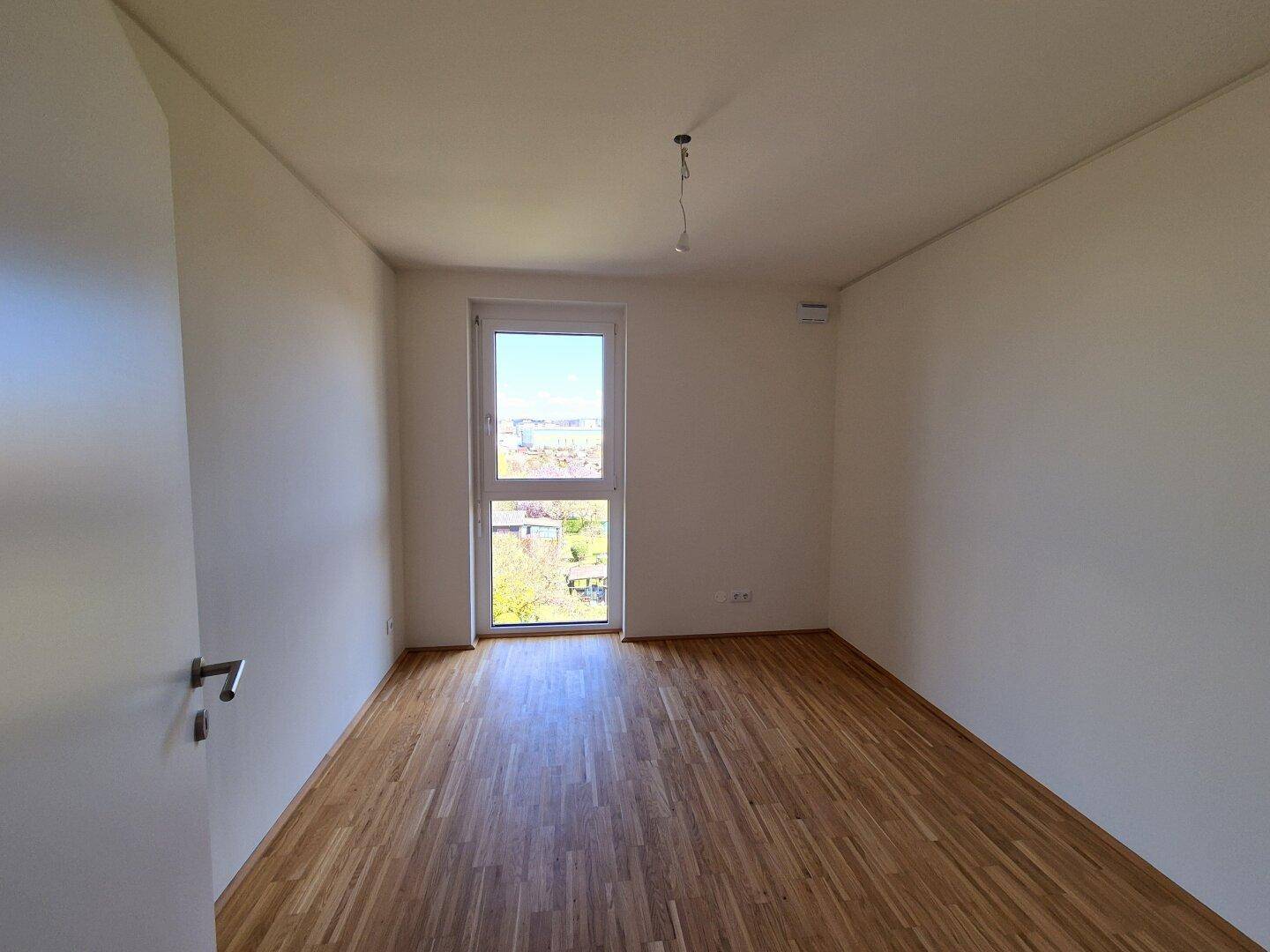 Helles Zimmer mit Parkettboden und einem Fenster, das einen Blick ins Grüne ermöglicht.