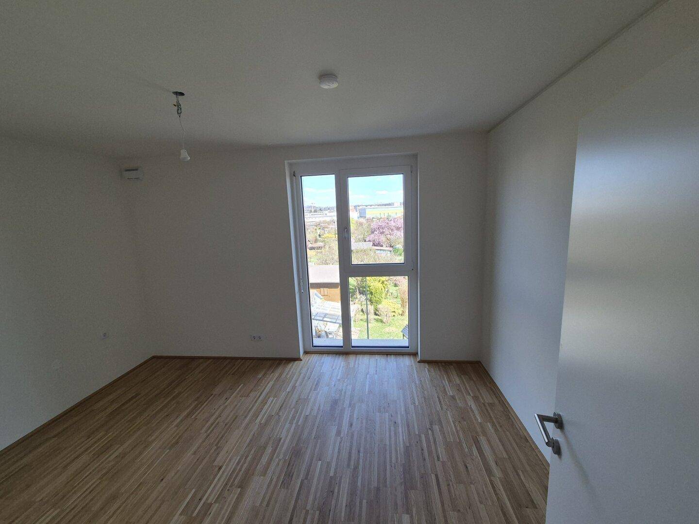 Helles Schlafzimmer mit Parkettboden und großem Fenster mit Blick auf die grüne Umgebung.