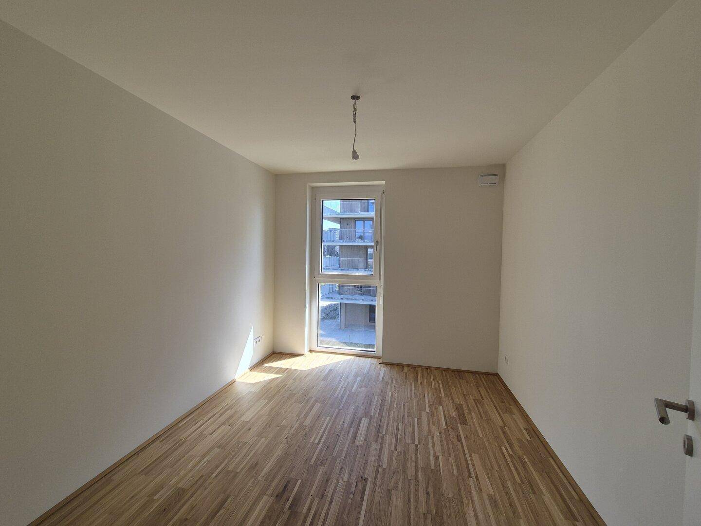 Leeres Schlafzimmer mit Parkettboden und großem Fenster, das viel Tageslicht hereinlässt.