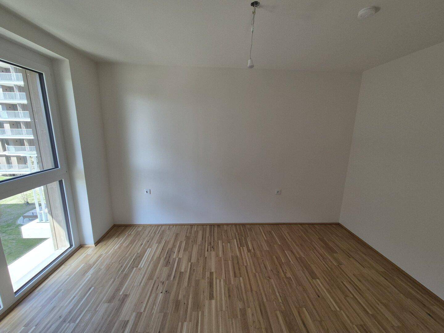 Helles Zimmer mit Parkettboden und großem Fenster, das einen Blick ins Grüne bietet.