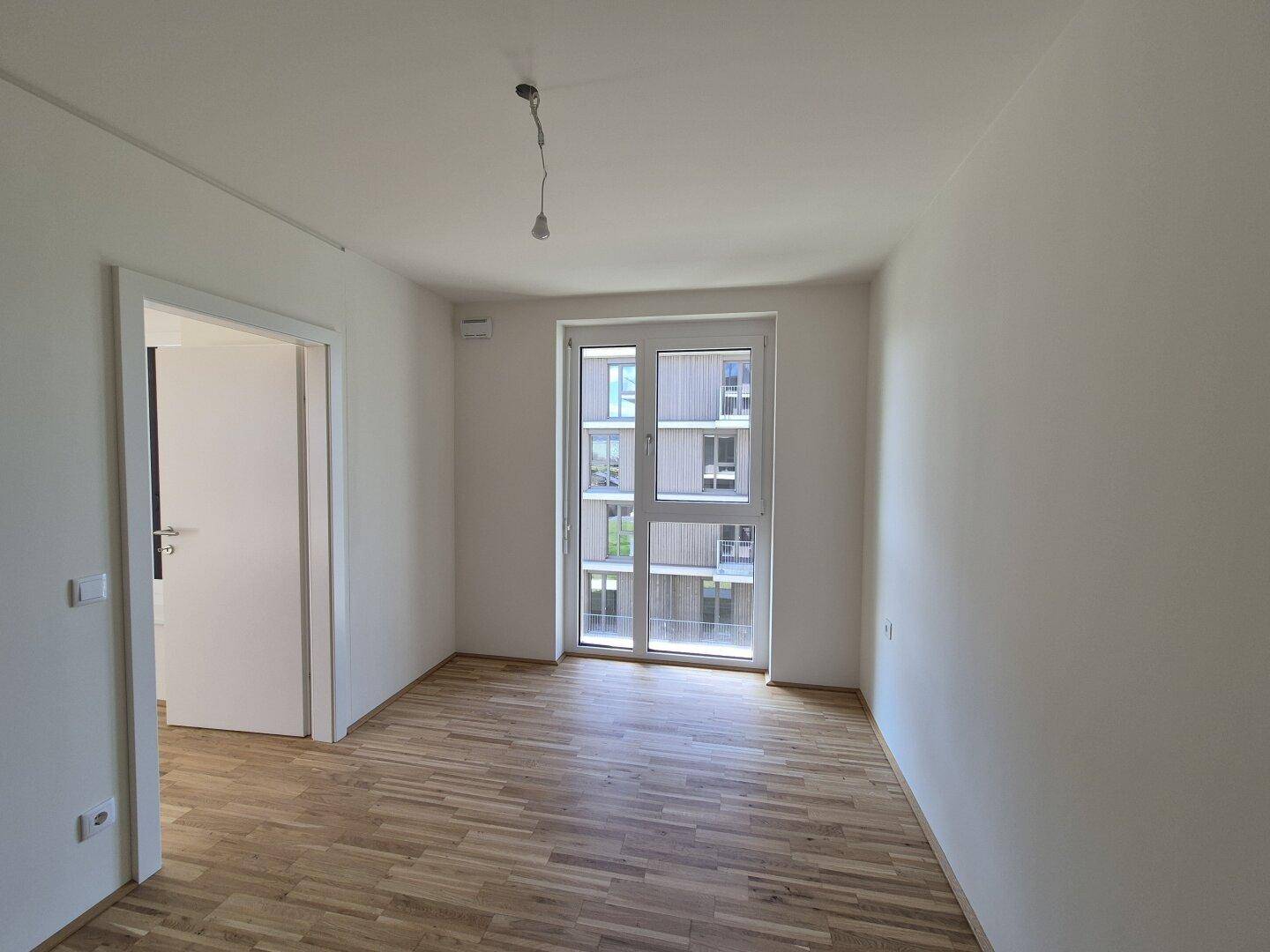 Leeres Schlafzimmer mit Parkettboden und Fenster mit Blick auf die umliegenden Gebäude.