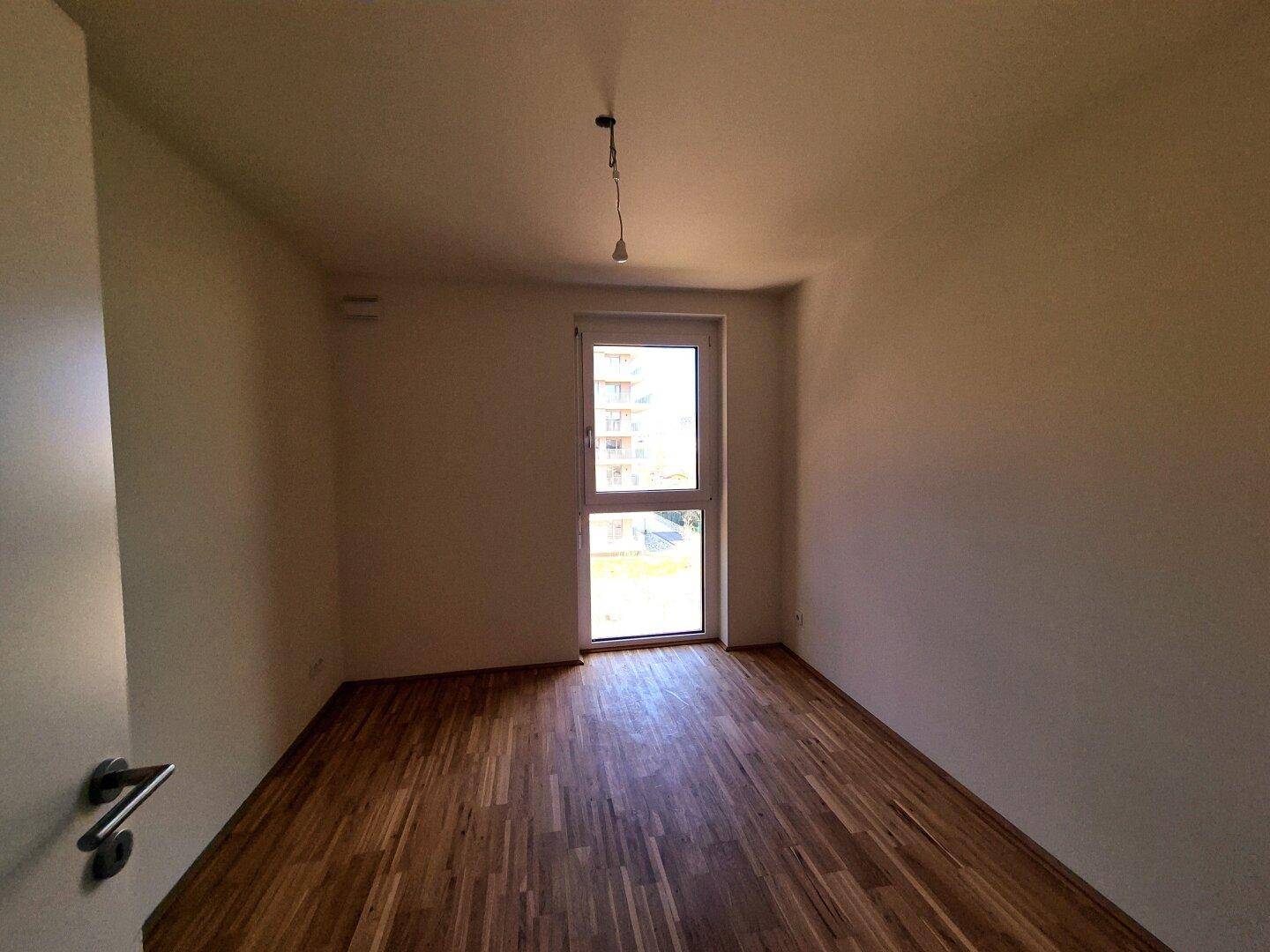 Schlafzimmer mit Holzparkettboden und großem Fenster, das viel Tageslicht hereinlässt.