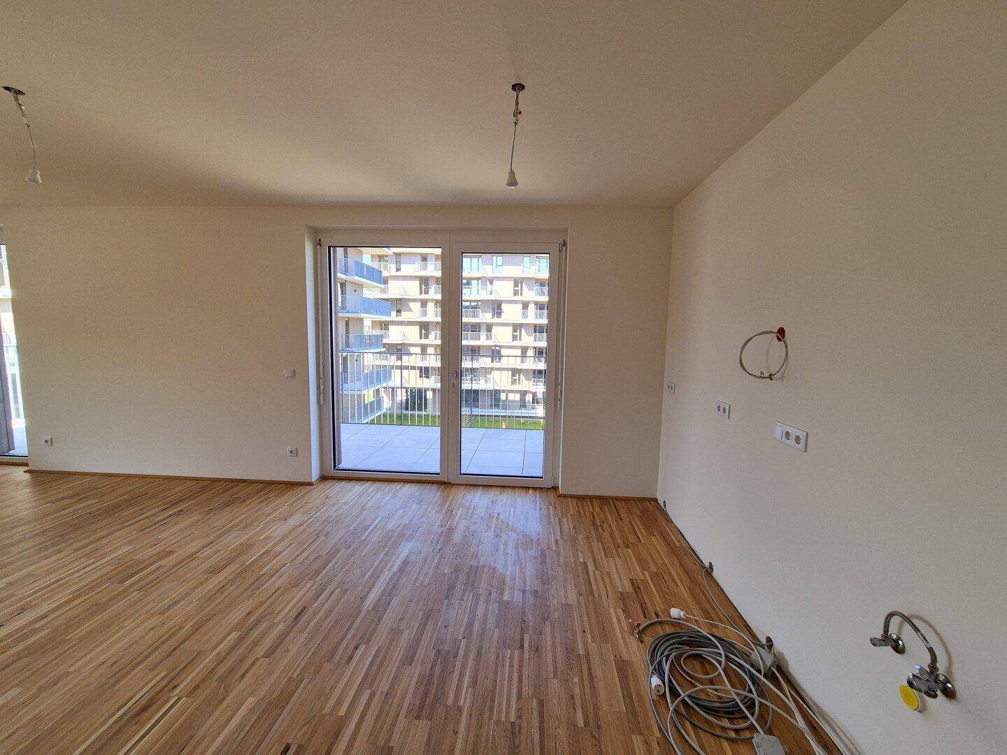 Geräumiger Wohnbereich mit Holzparkettboden und Zugang zu einem Balkon, vorbereitet für eine Küche.
