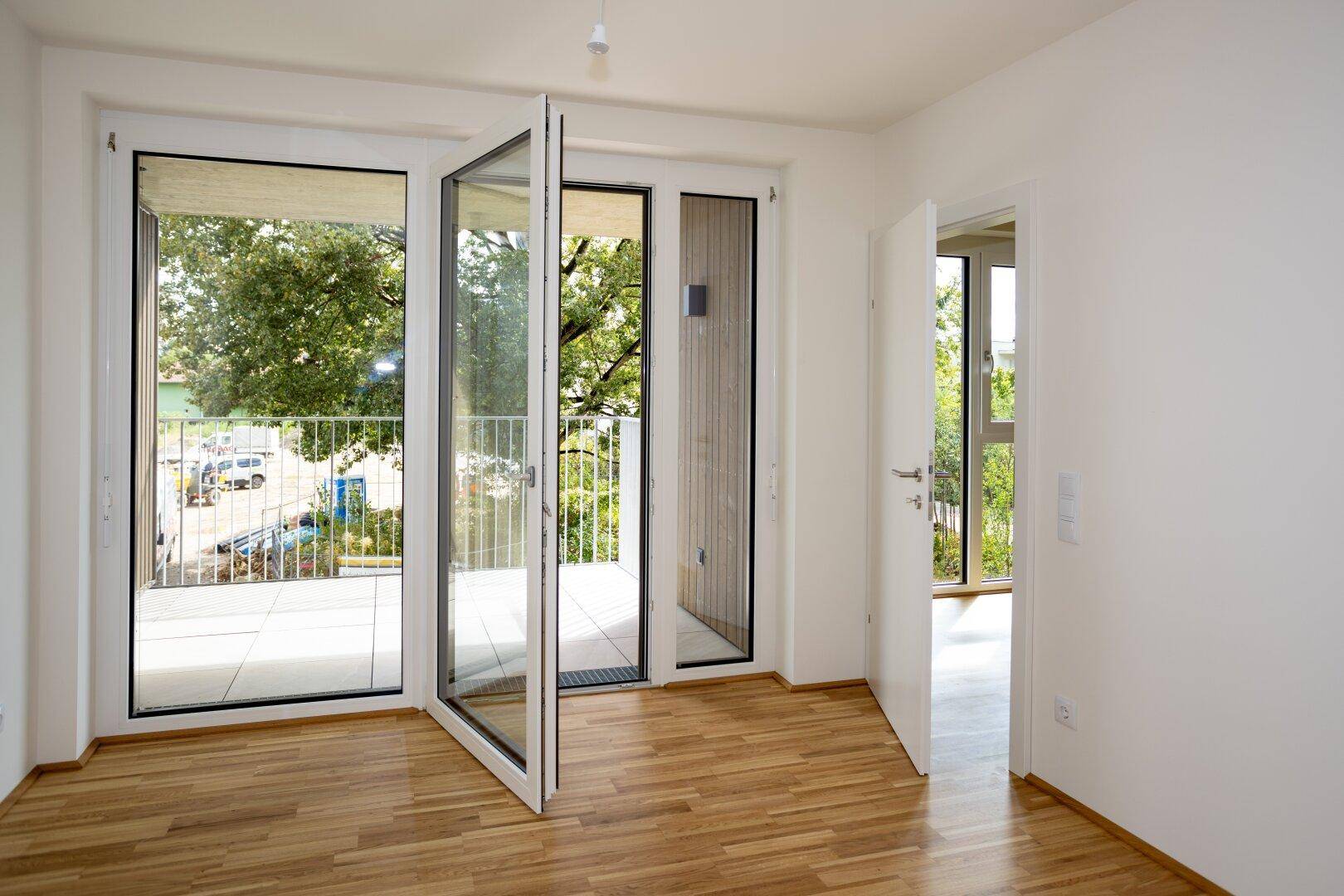 Heller, leerer Raum mit Parkettboden und großen Fenstertüren zum Balkon.