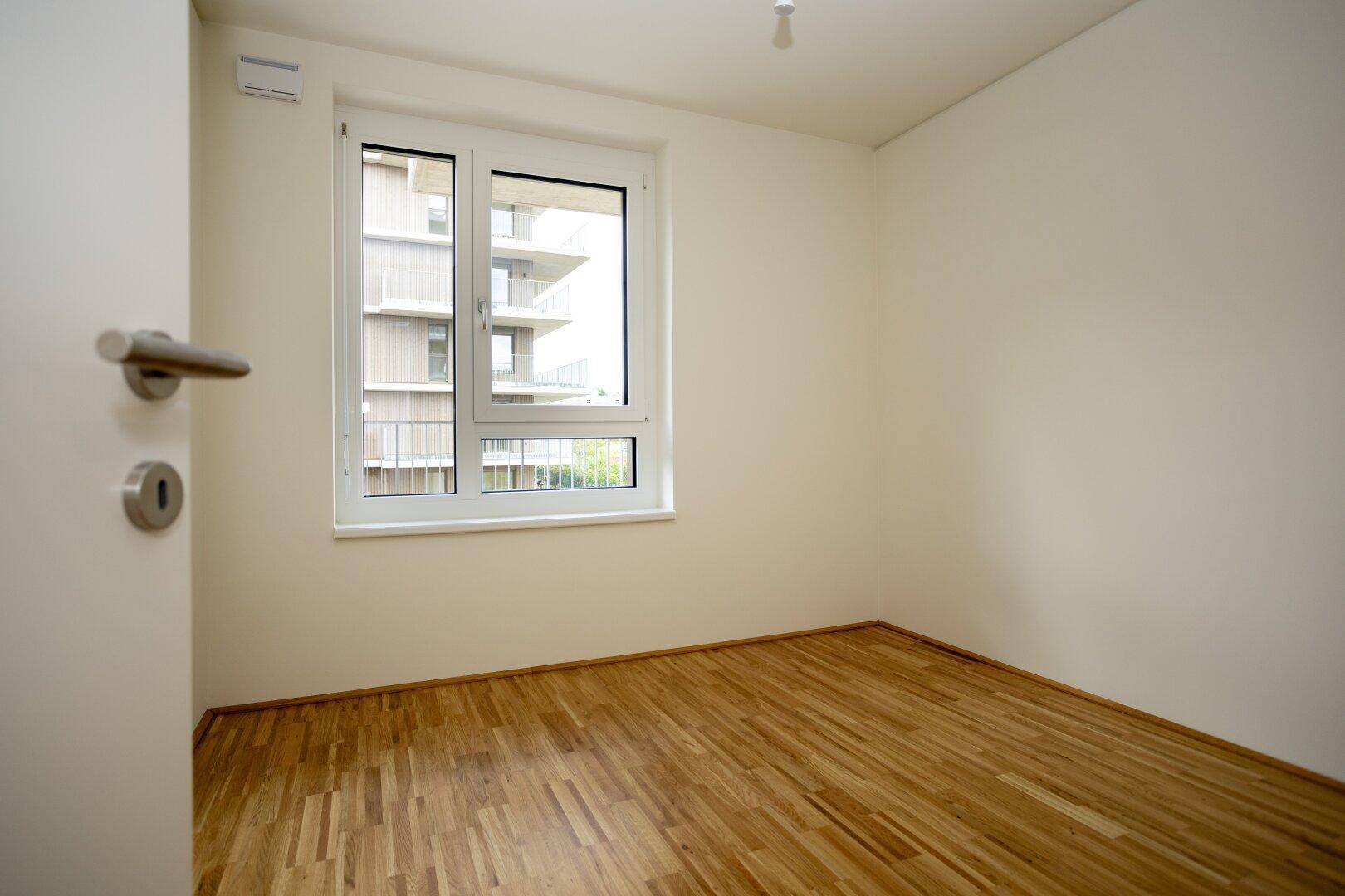 Helles Schlafzimmer mit Parkettboden und Fensterblick auf die moderne Wohnanlage.