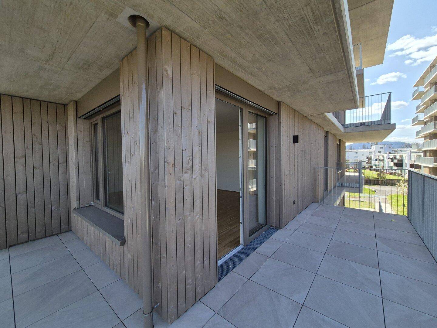 Heller Balkon mit Fliesenboden und Holzverkleidung an der Fassade, mit Zugang zu den Innenräumen.
