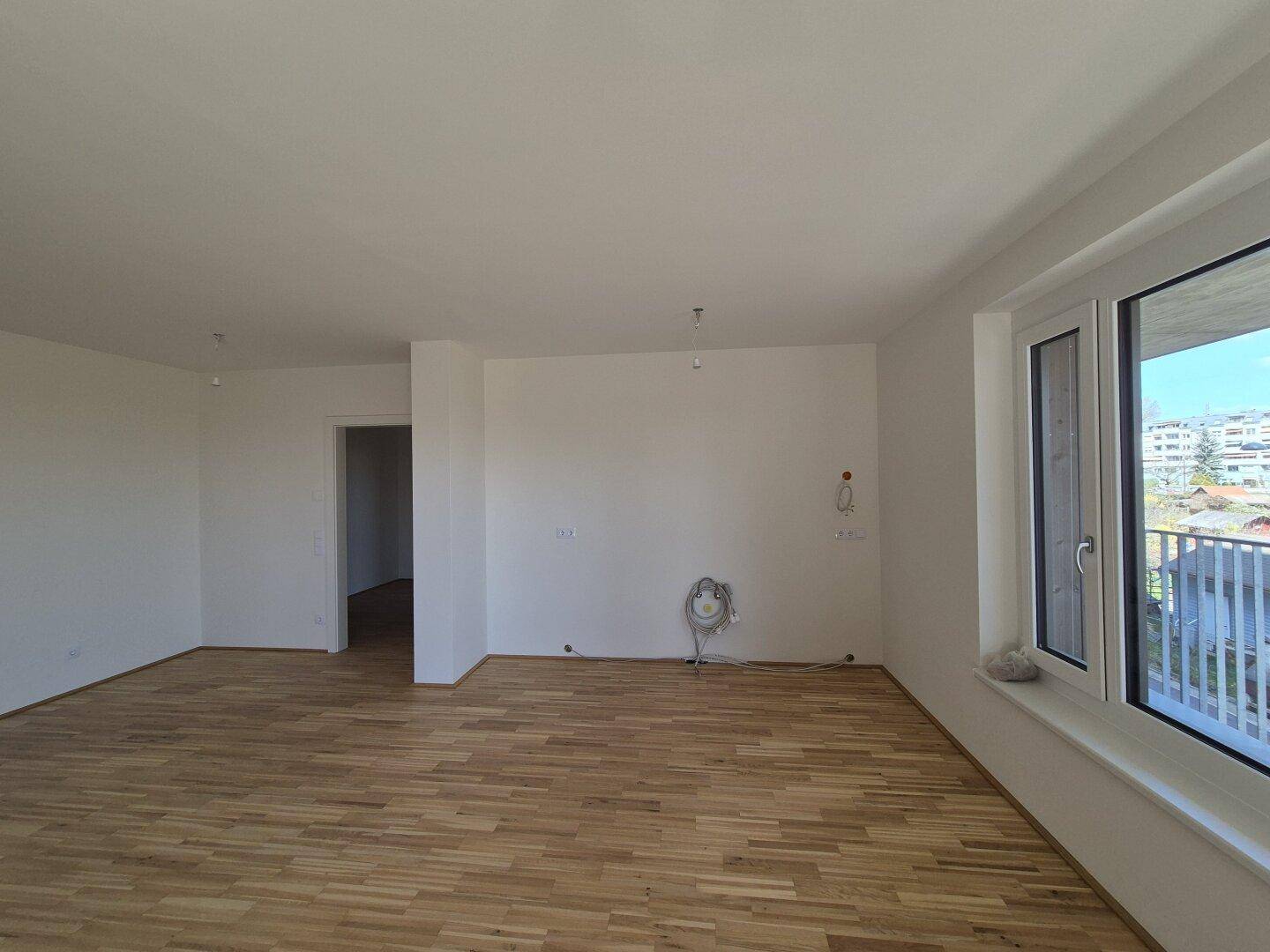 Heller Wohnbereich mit Parkettboden und großem Fenster, das viel Tageslicht hereinlässt.