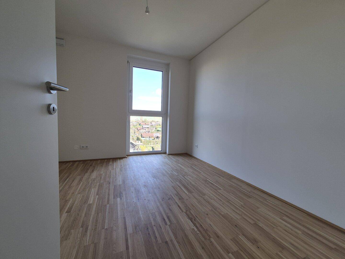 Schlafzimmer mit Parkettboden und großem Fenster, das viel Tageslicht hereinlässt.