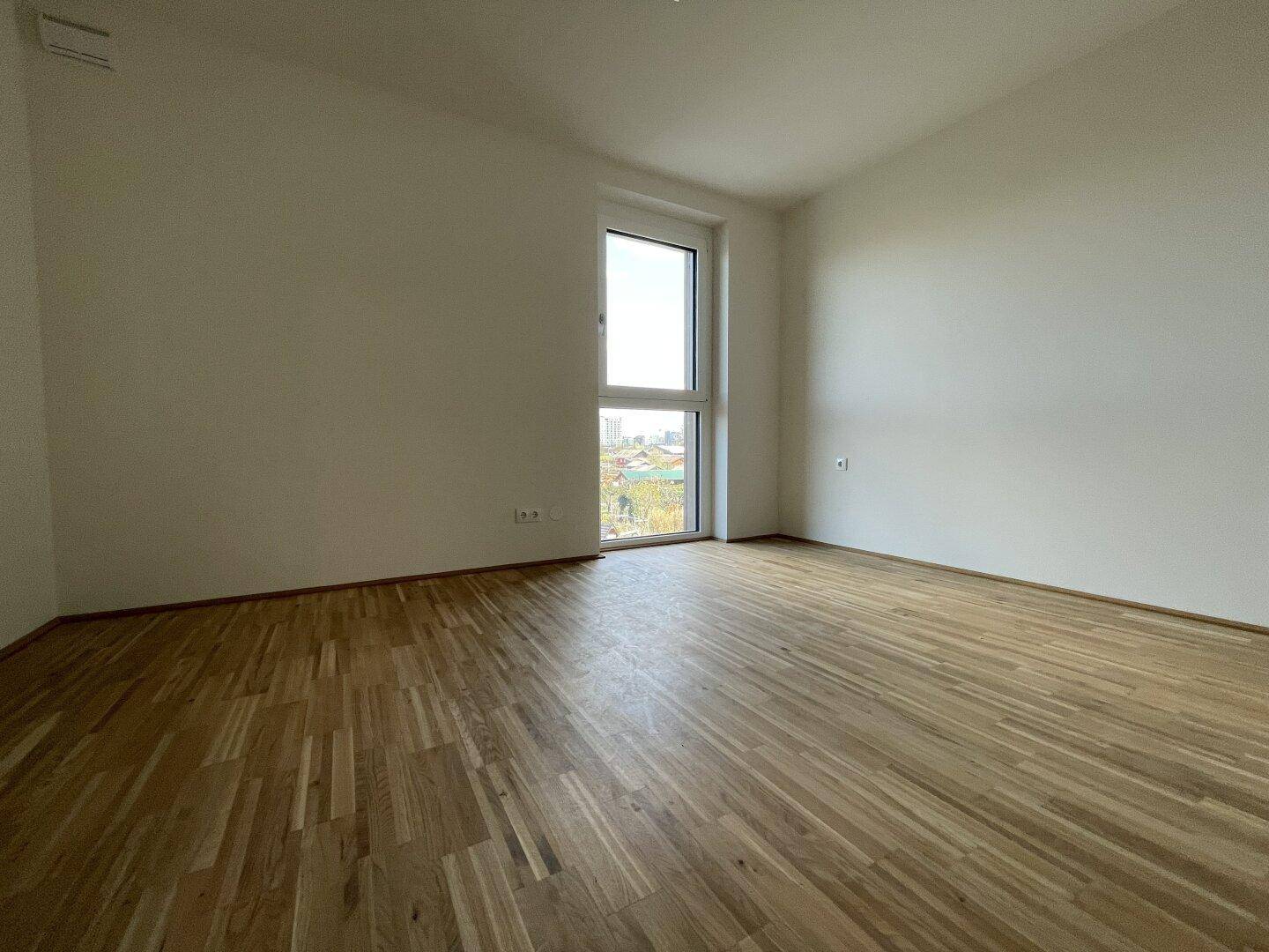 Helles Schlafzimmer mit Parkettboden und großem Fenster, bietet einen Ausblick auf die umliegenden Gebäude.