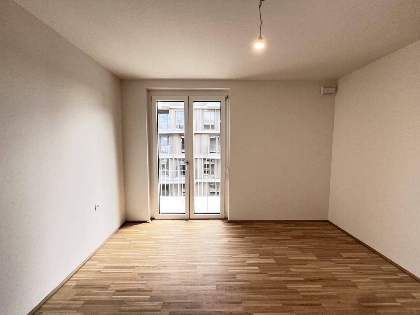 Heller, leerer Raum mit hellem Parkettboden und Zugang zu einem Balkon durch eine große Glastür.