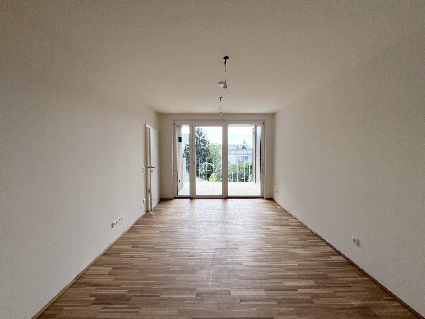 Geräumiges, leeres Zimmer mit Holzboden und großen Fenstern, die viel Tageslicht hereinlassen.
