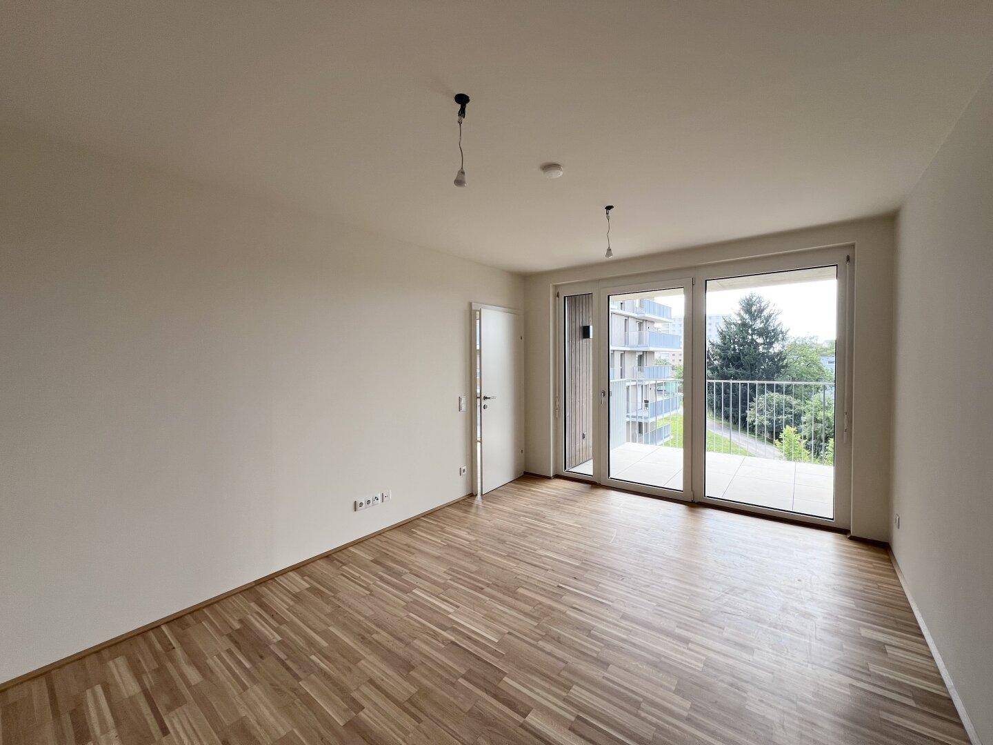 Leeres Zimmer mit Parkettboden und Balkonzugang, bietet einen Blick auf die umliegenden Gebäude.