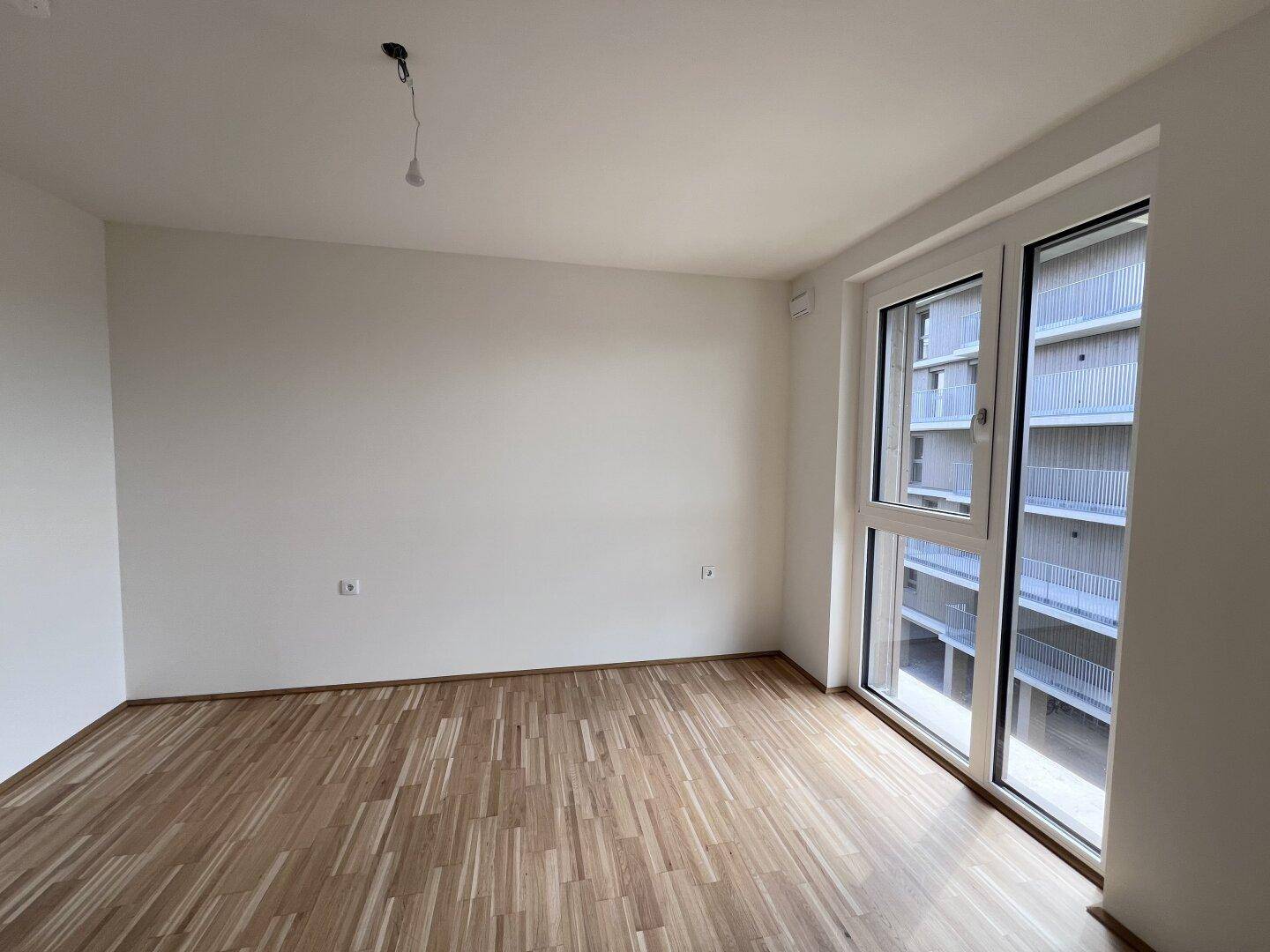 Helles Schlafzimmer mit Parkettboden und großem Fenster, das viel Tageslicht hereinlässt.