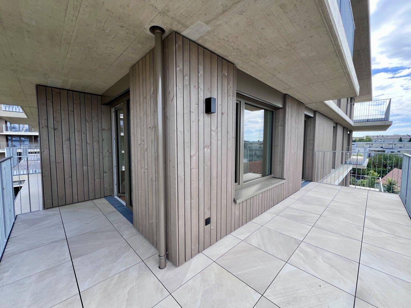 Moderner Balkon mit Holzverkleidung und Fliesenboden, umgeben von anderen Gebäuden der Anlage.