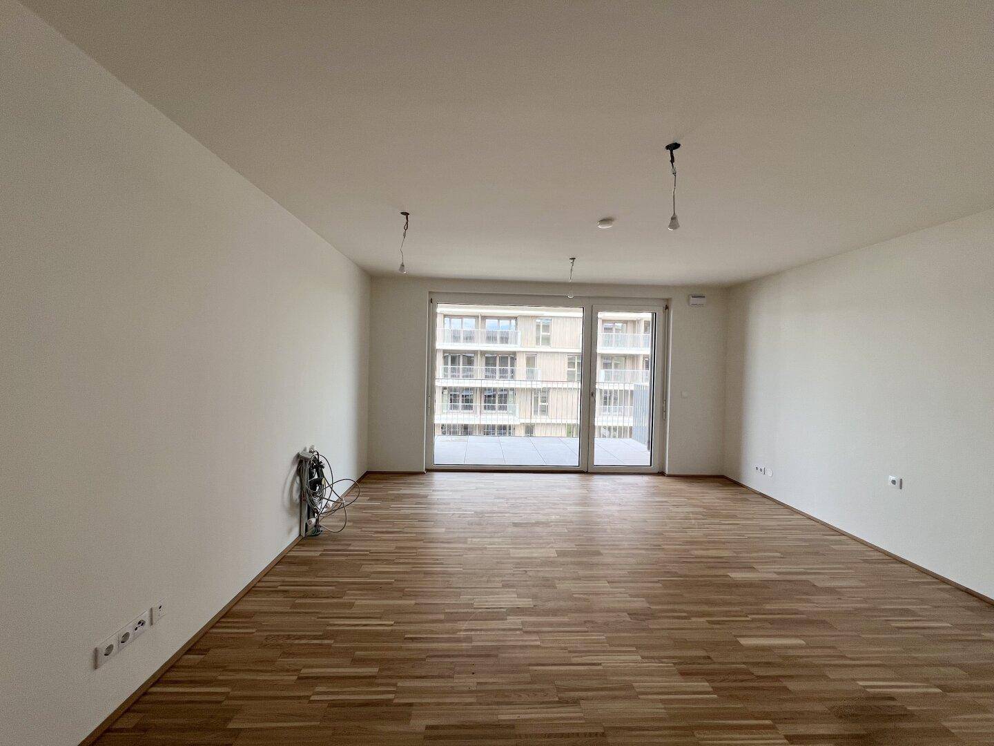 Heller, leerer Wohnraum mit Parkettboden und großen Fenstern, die viel Tageslicht hereinlassen.
