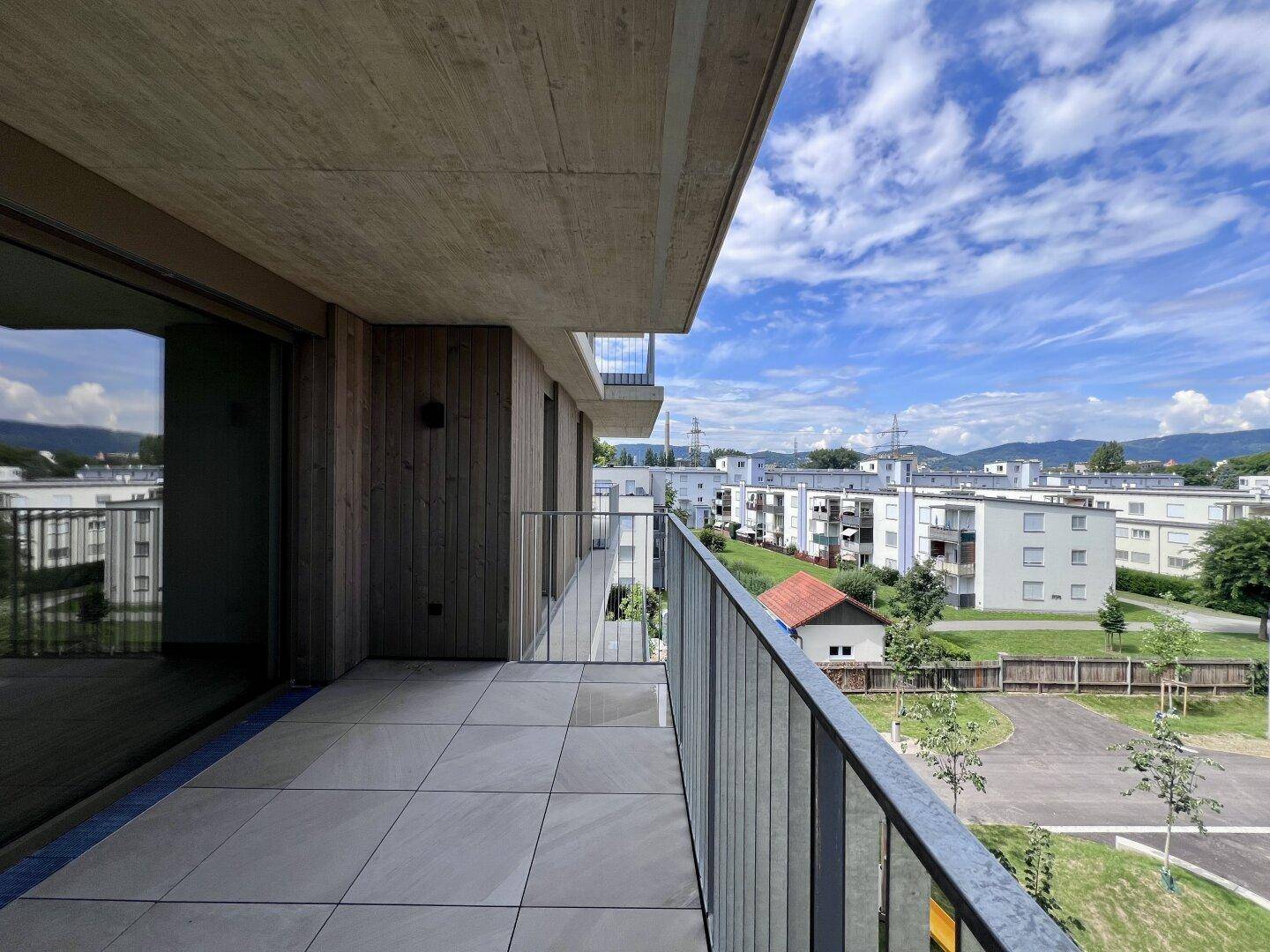 Langer Balkon mit Fliesenboden und Glasgeländer, der eine weite Aussicht auf die Wohngegend ermöglicht.