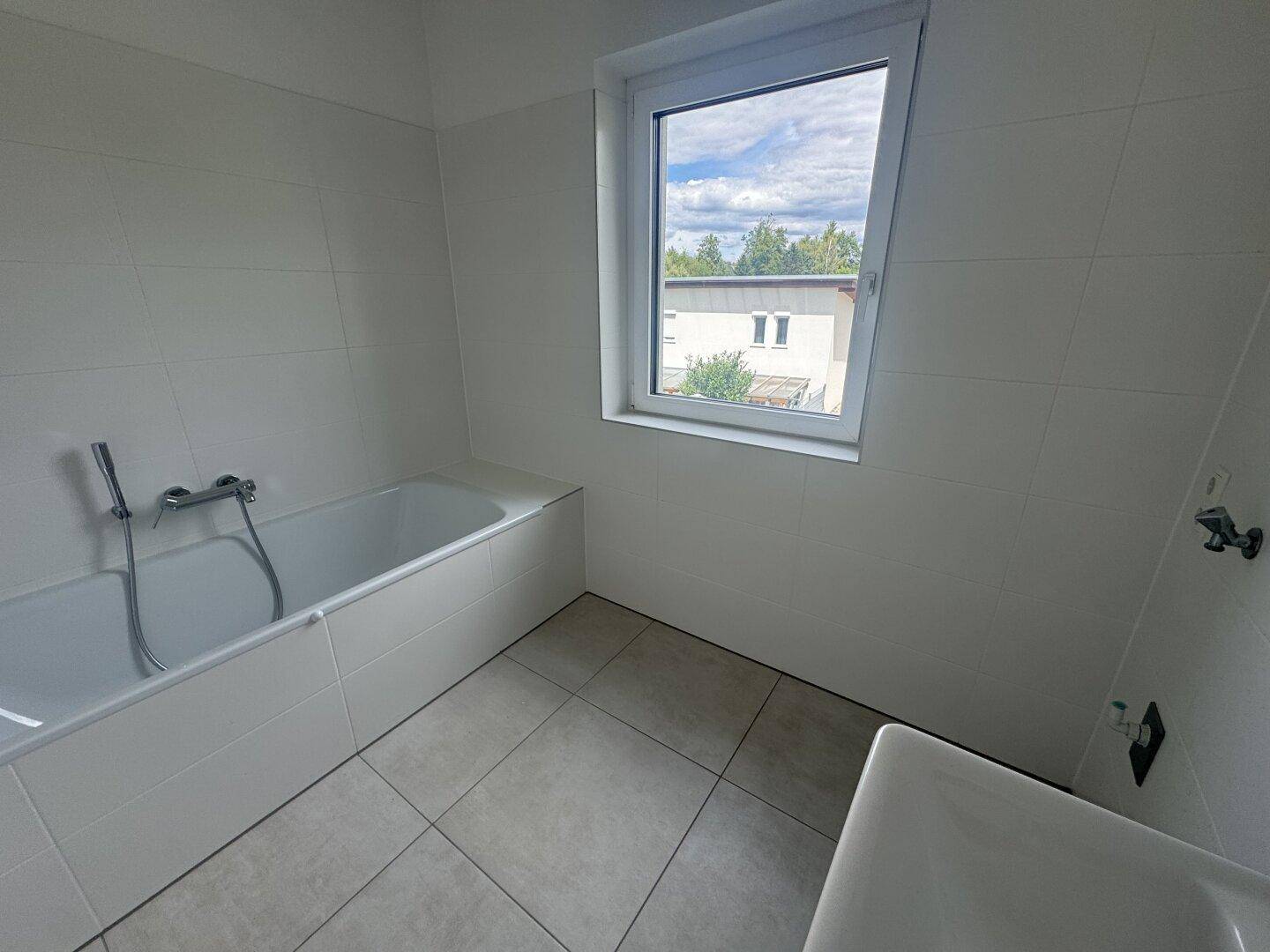 Helles Badezimmer mit Badewanne, Waschbecken, Fenster und modernen weißen Fliesen.