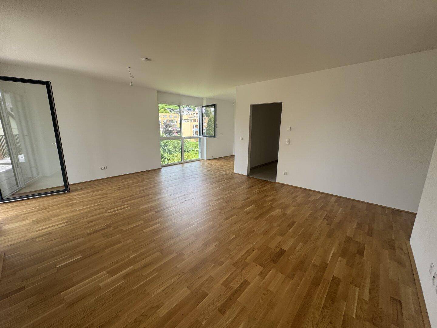 Geräumiges Wohnzimmer mit hellem Parkettboden, weißen Wänden und großen Fenstern für viel Tageslicht.