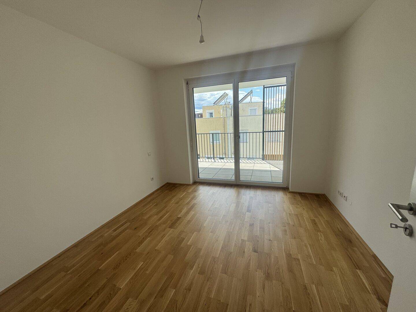 Schlafzimmer mit Parkettboden, weißen Wänden und direktem Zugang zu einem sonnigen Balkon.