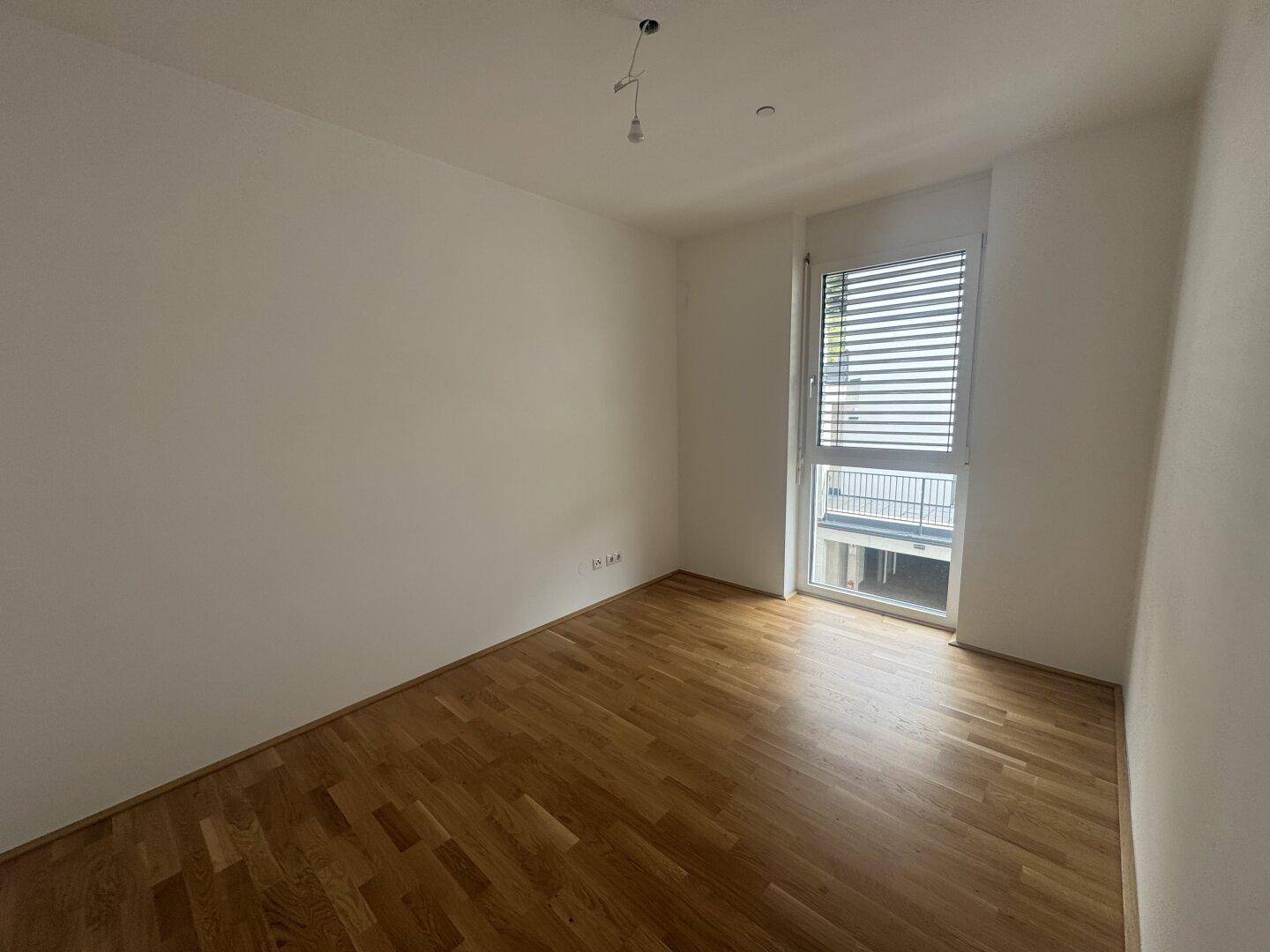 Ruhiges Schlafzimmer mit Parkettboden, weißen Wänden und einem Fenster mit Jalousien.
