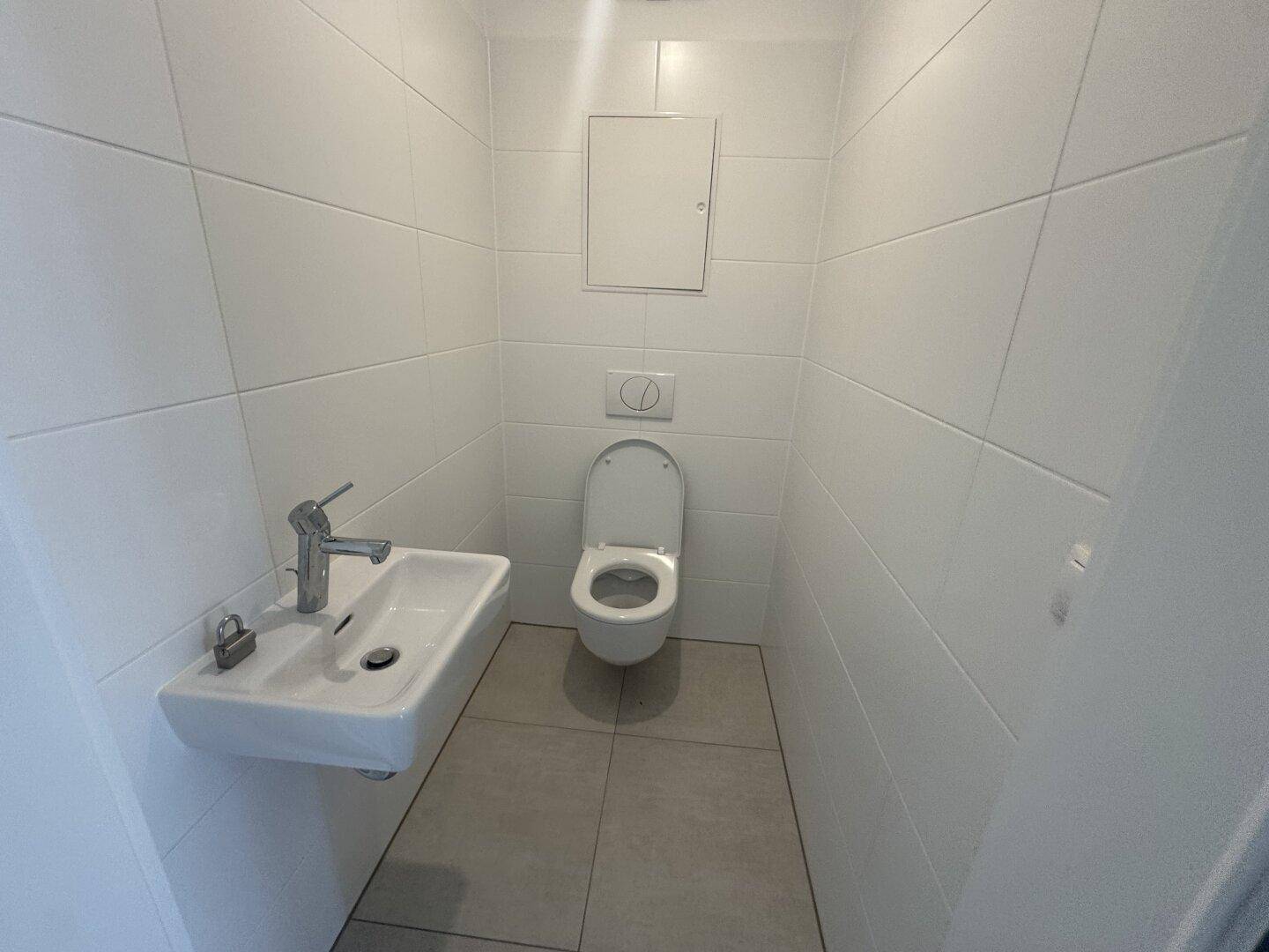 Modernes Gäste-WC mit Wand-WC, kleinem Waschbecken und hellen Fliesen an Wänden und Boden.