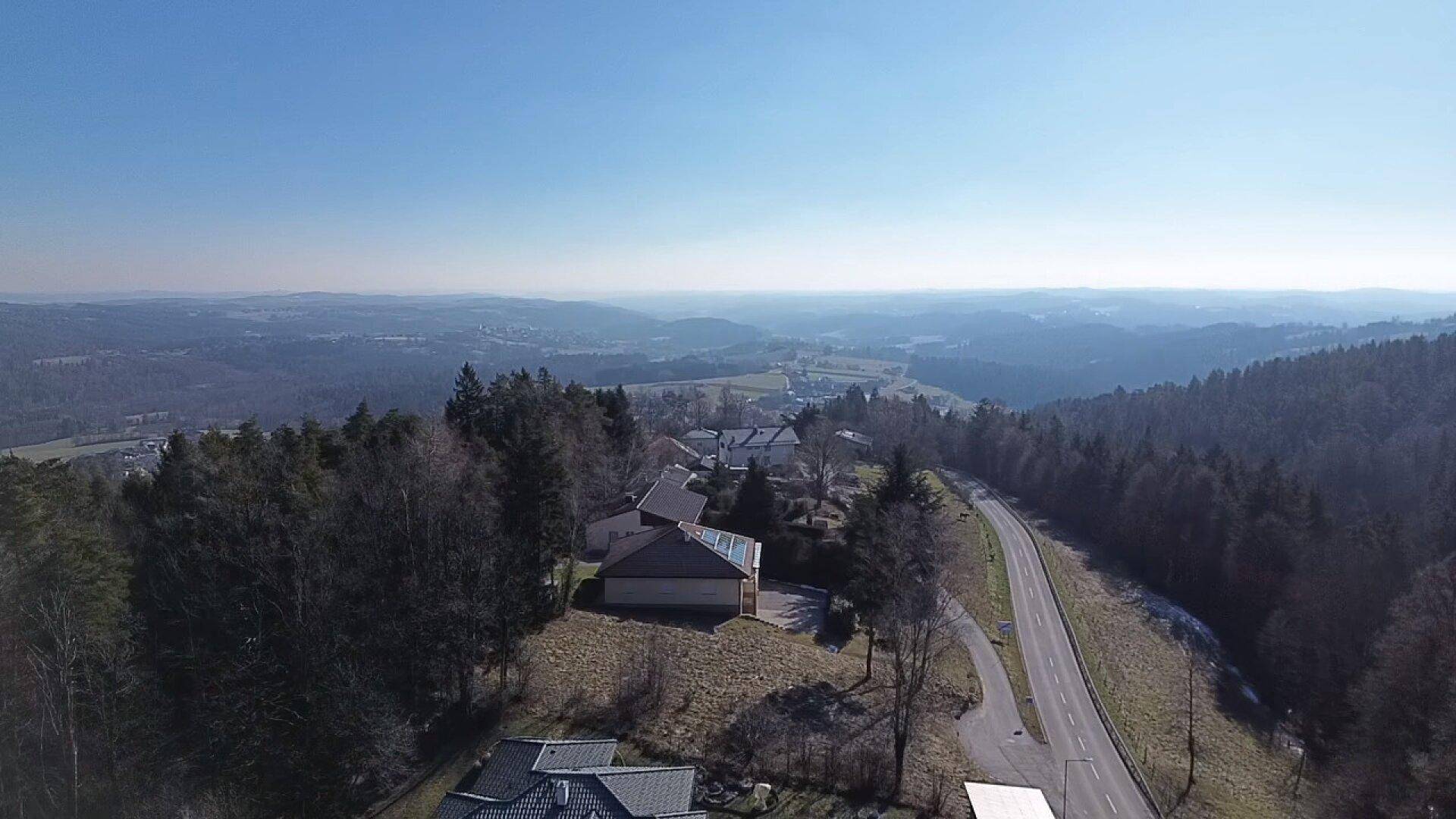 Weitläufige Panoramaansicht einer hügeligen Landschaft mit Wald, Häusern und einer Straße unter blauem Himmel.