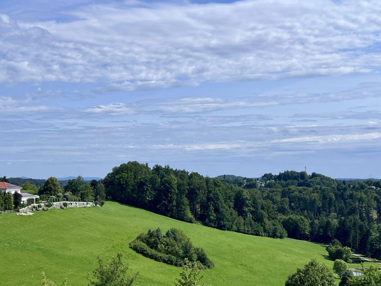 Idyllisches Haus eingebettet in eine weitläufige grüne Hügellandschaft mit dichten Wäldern und einem malerischen Fernblick unter blauem Himmel.