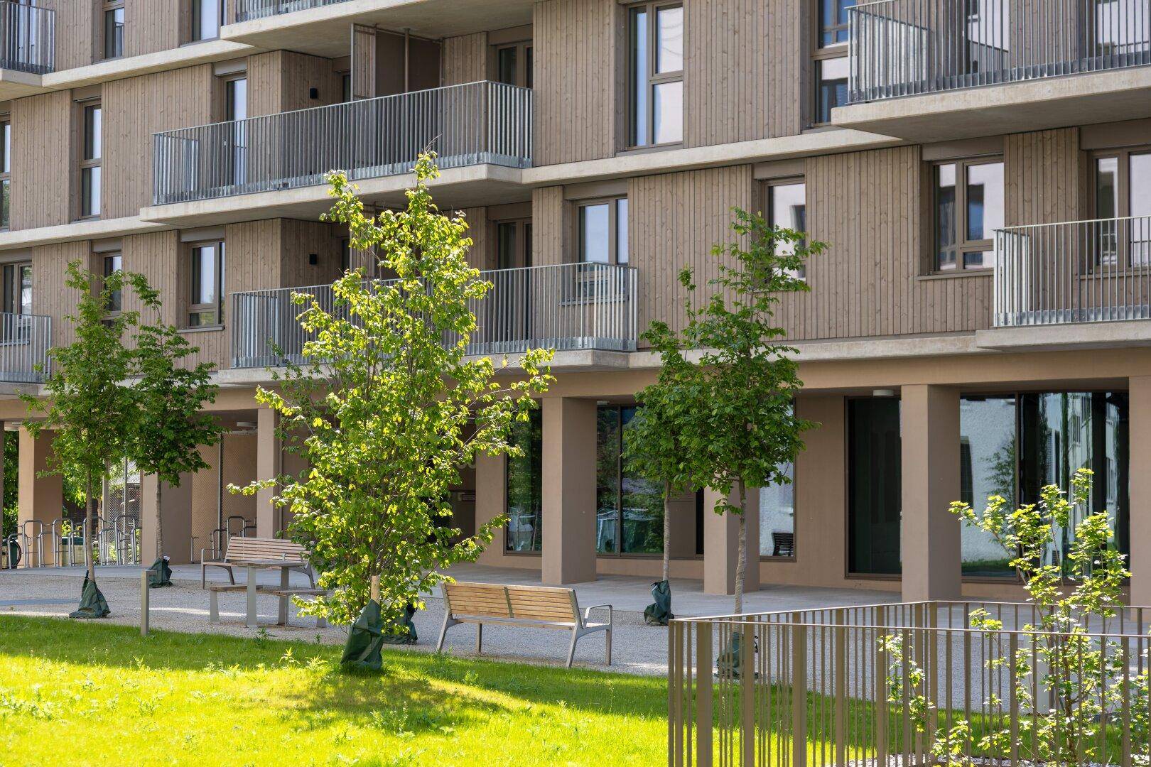 Außenansicht eines modernen Wohngebäudes mit Holzfassade, Balkonen und Sitzbänken im begrünten Außenbereich.