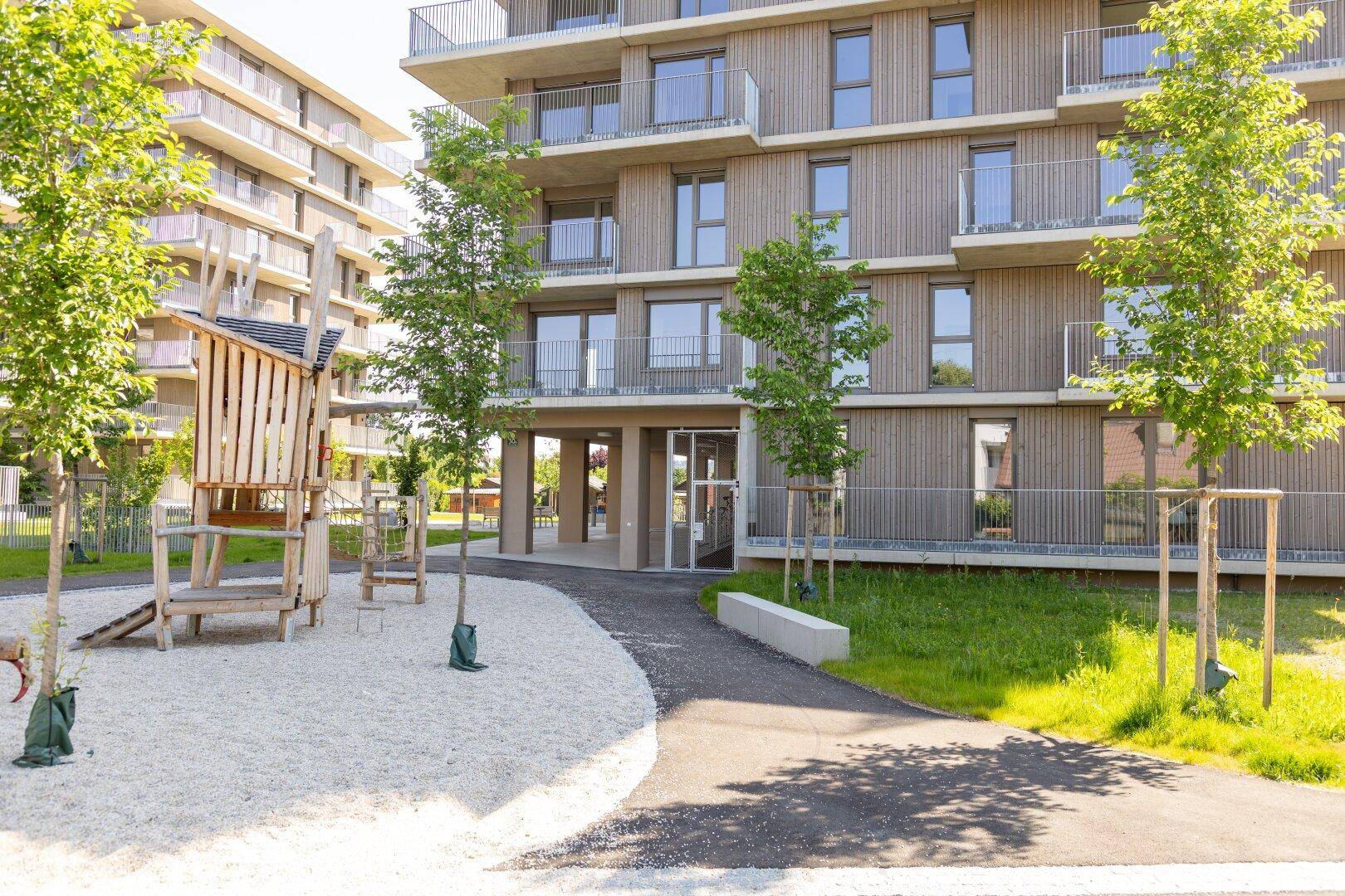 Moderner Wohnkomplex mit Holzfassade, Balkonen und einem Spielplatz mit Sandfläche und Holzspielgeräten.