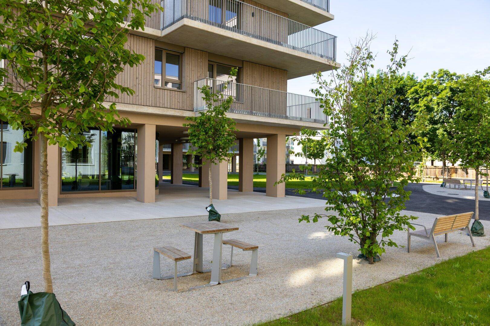 Außenbereich eines modernen Wohnkomplexes mit Sitzgelegenheiten, Bäumen und gepflegten Kiesflächen.