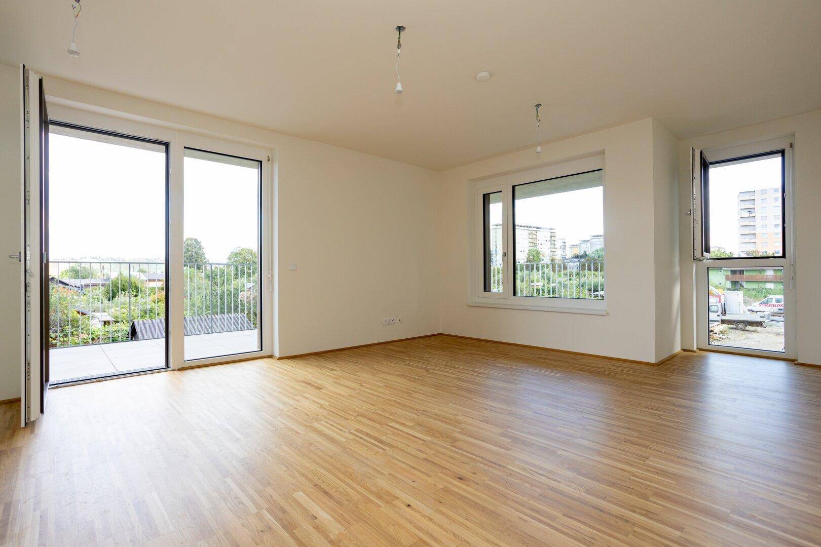 Geräumiges Wohnzimmer mit Holzboden, großen Fenstern und Zugang zum Balkon mit Weitblick.
